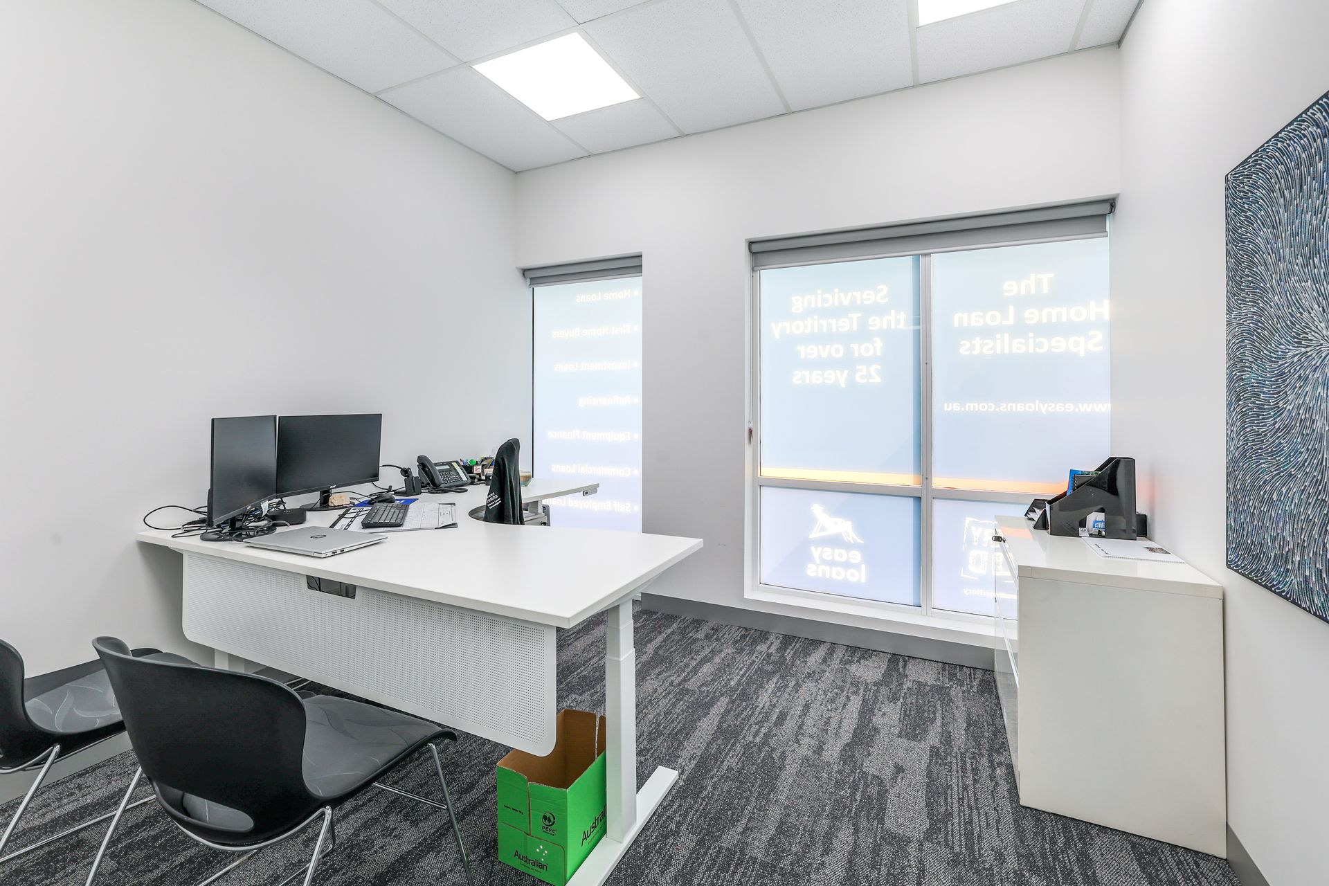 Office with white desk, dual monitors, gray carpet, and a window with text; a black chair and a small side table.