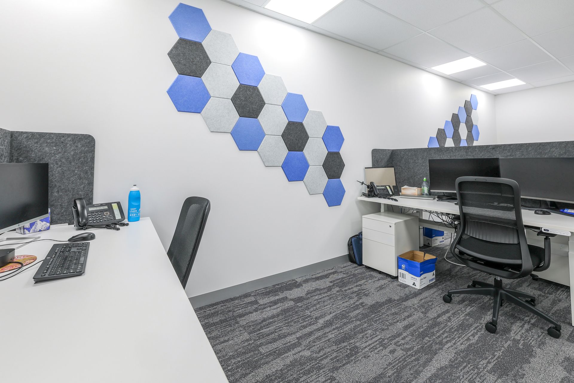 Office space with white desks, black chairs, gray carpet, and blue/gray honeycomb wall decor.
