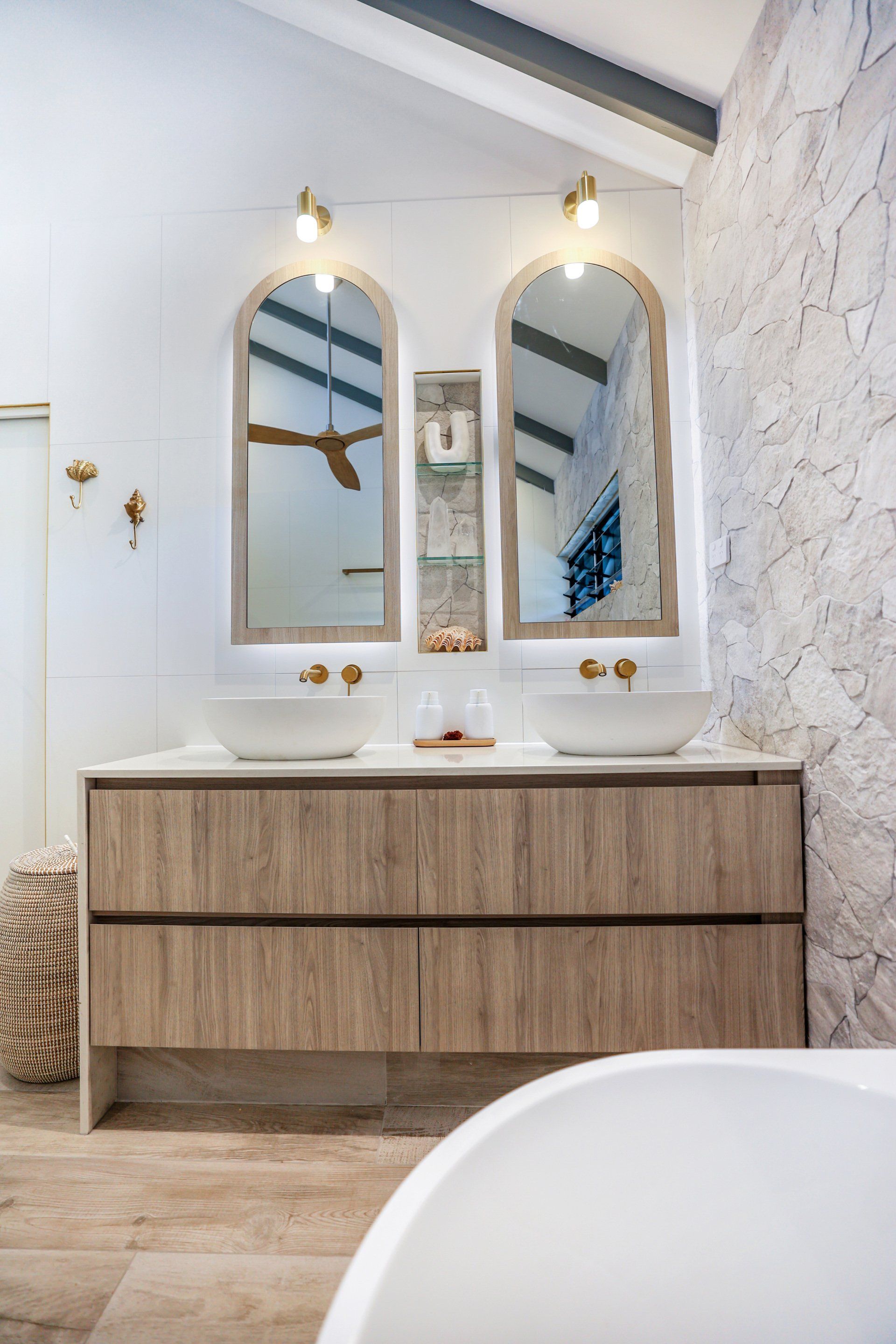 Bathroom with double sinks, arched mirrors, and textured wall. Light wood vanity with white sinks, gold accents.