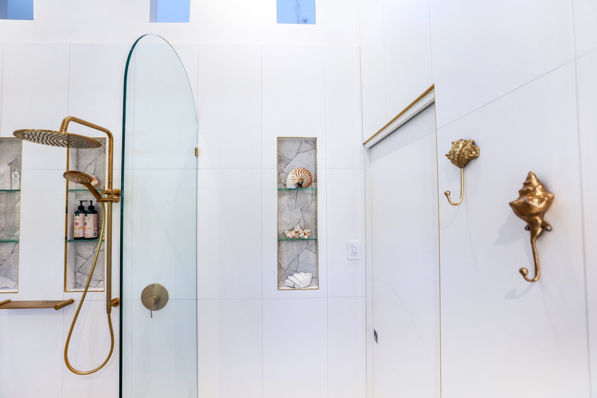 White tiled bathroom with a gold shower, glass door, seashell hooks, and built-in shelves.