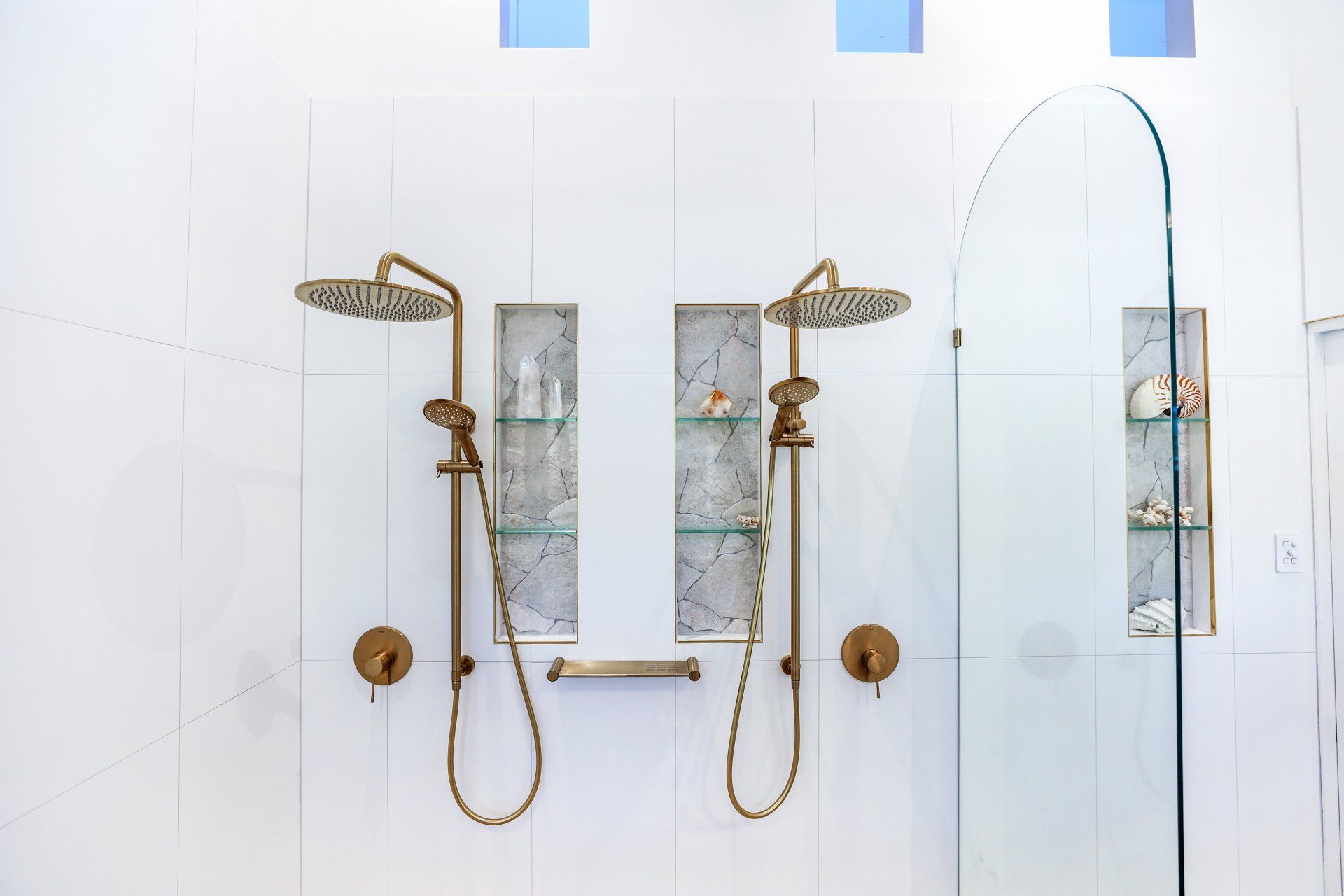 Two gold showerheads with hand showers in a white tile bathroom, with glass door and shelves.