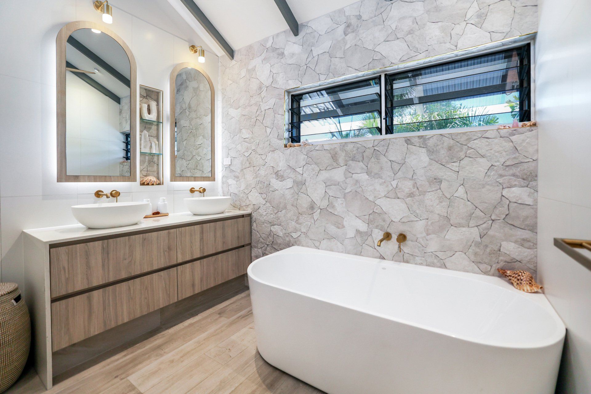 Bathroom with stone wall, double vanity, arched mirrors, and a freestanding bathtub.