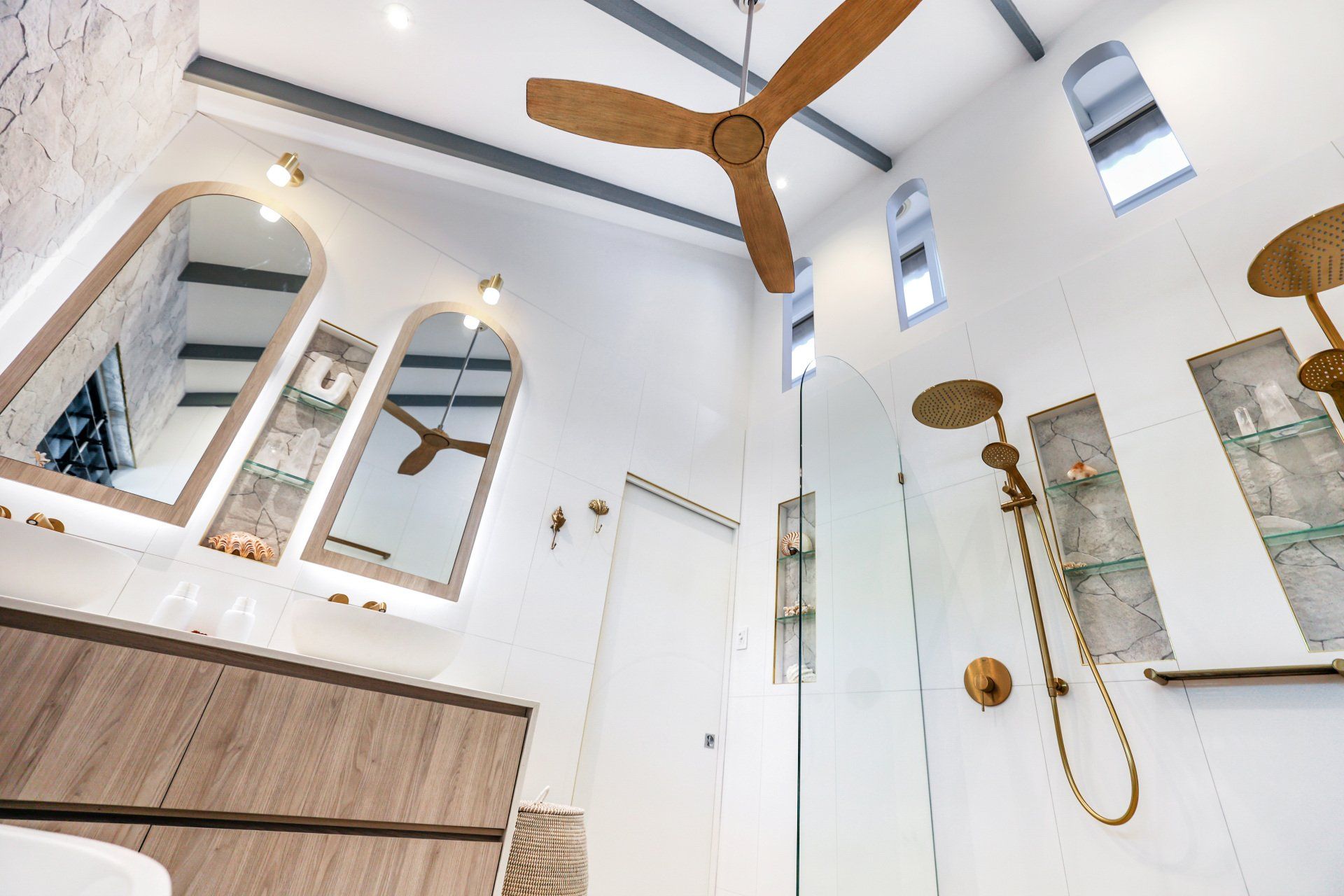Modern bathroom with wood ceiling fan, gold fixtures, arched mirrors, and a glass shower.