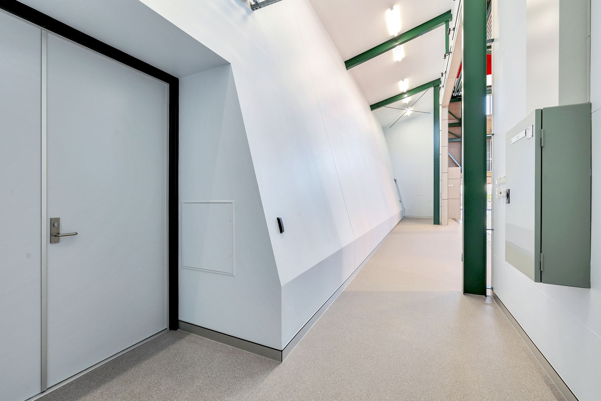 Hallway with white walls and angled ceiling, door on left, green beams, gray floor, gray box.