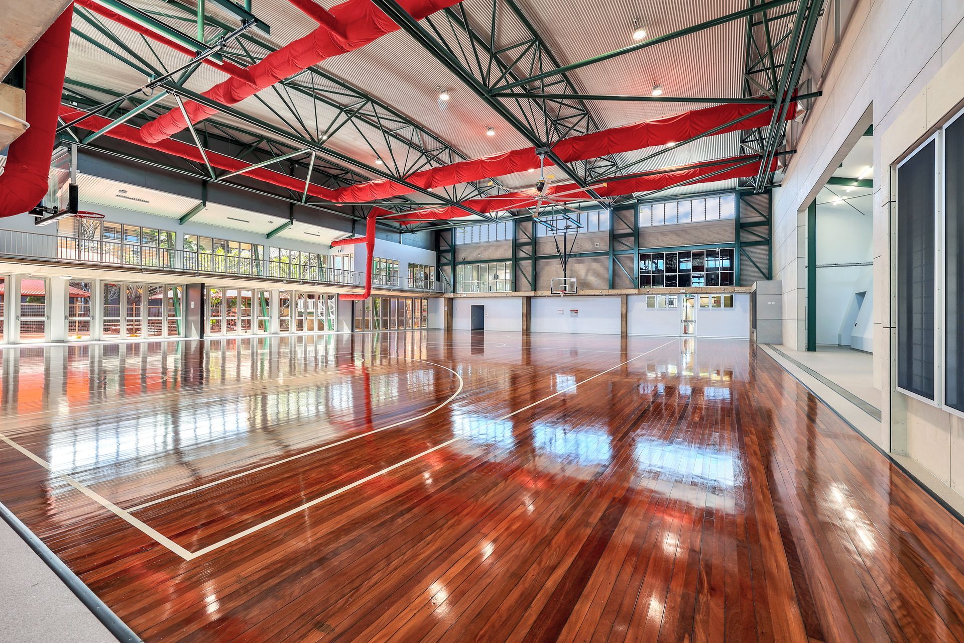 Wooden basketball court inside gymnasium with red accents, windows and glass panels.