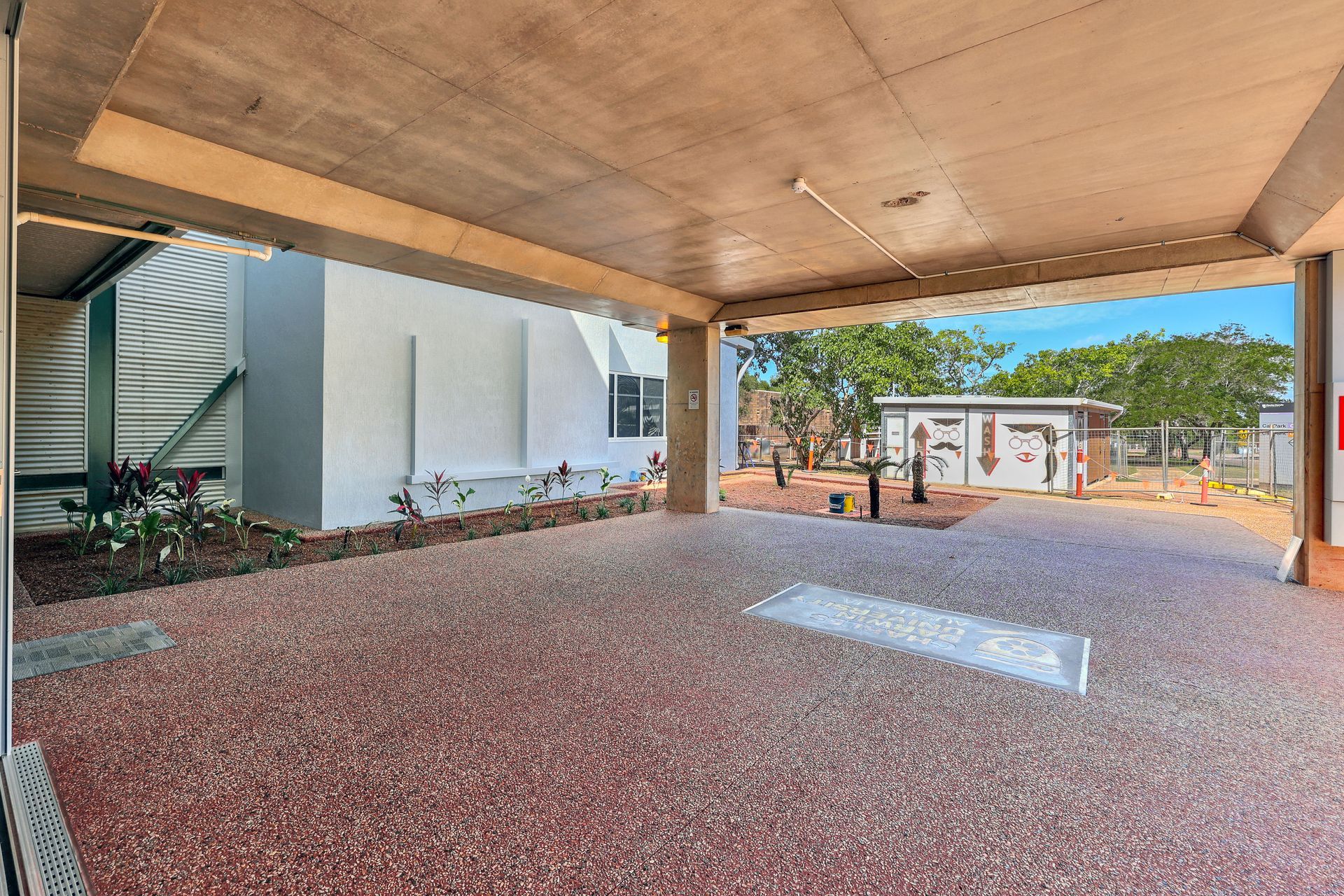 Covered outdoor area, red pebbled floor, leading to open-air space with buildings and trees visible in the distance.
