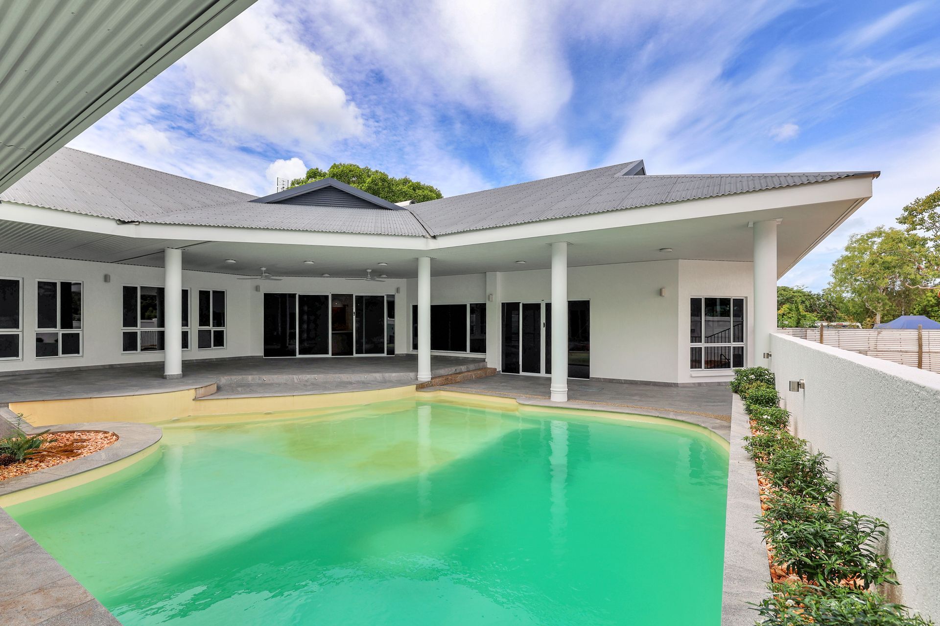 Pool in courtyard of a white house with a gray roof under a blue sky.