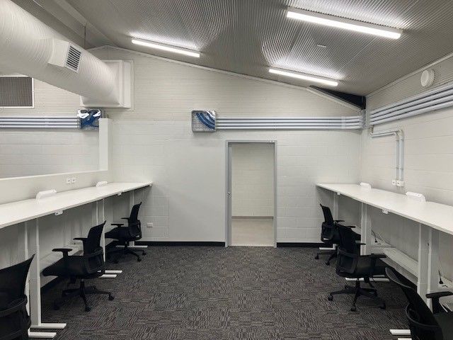 An empty office space with long desks, chairs, and an open doorway. White walls and ceiling, grey carpet.