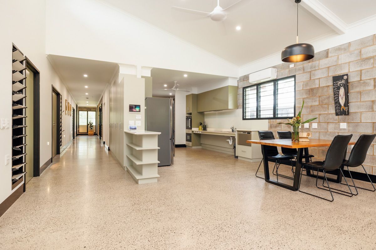 Open-plan kitchen and dining area. Beige speckled floor, brick wall. Dining table with black chairs. Kitchen cabinets are green.