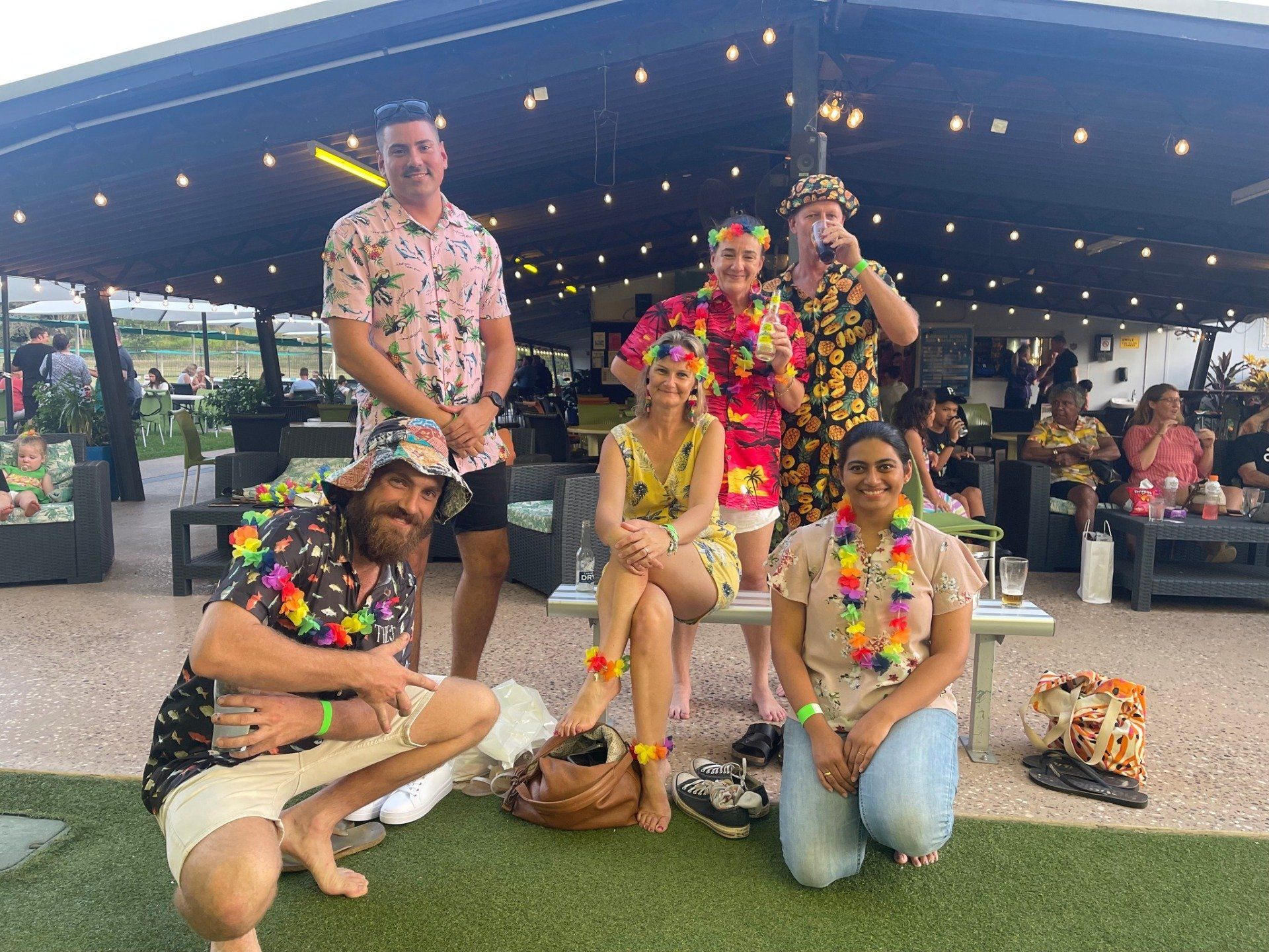 Group of people in Hawaiian shirts and leis posing outdoors under string lights.