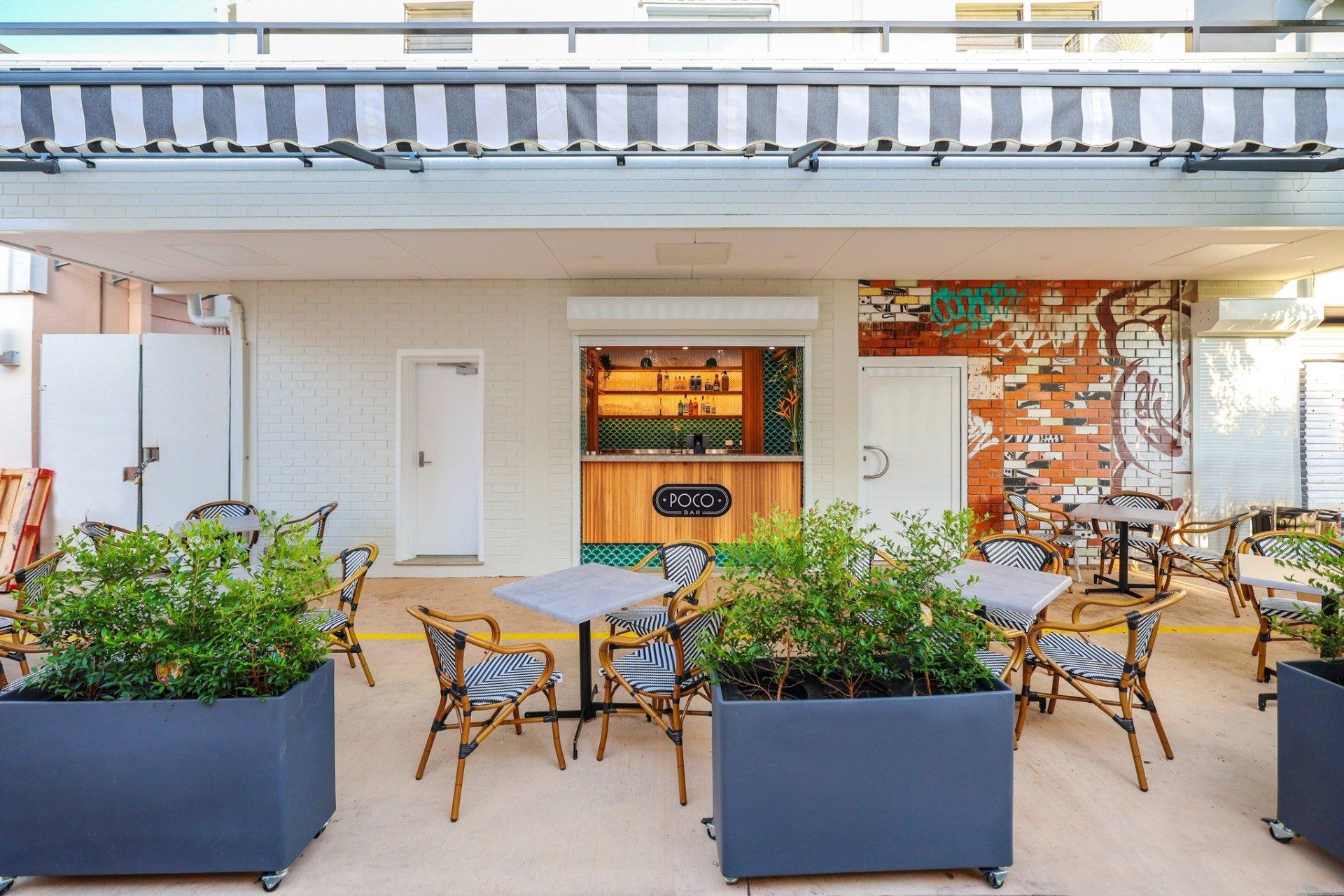 Outdoor cafe with tables, chairs, and potted plants under a striped awning. A bar is visible in the background.
