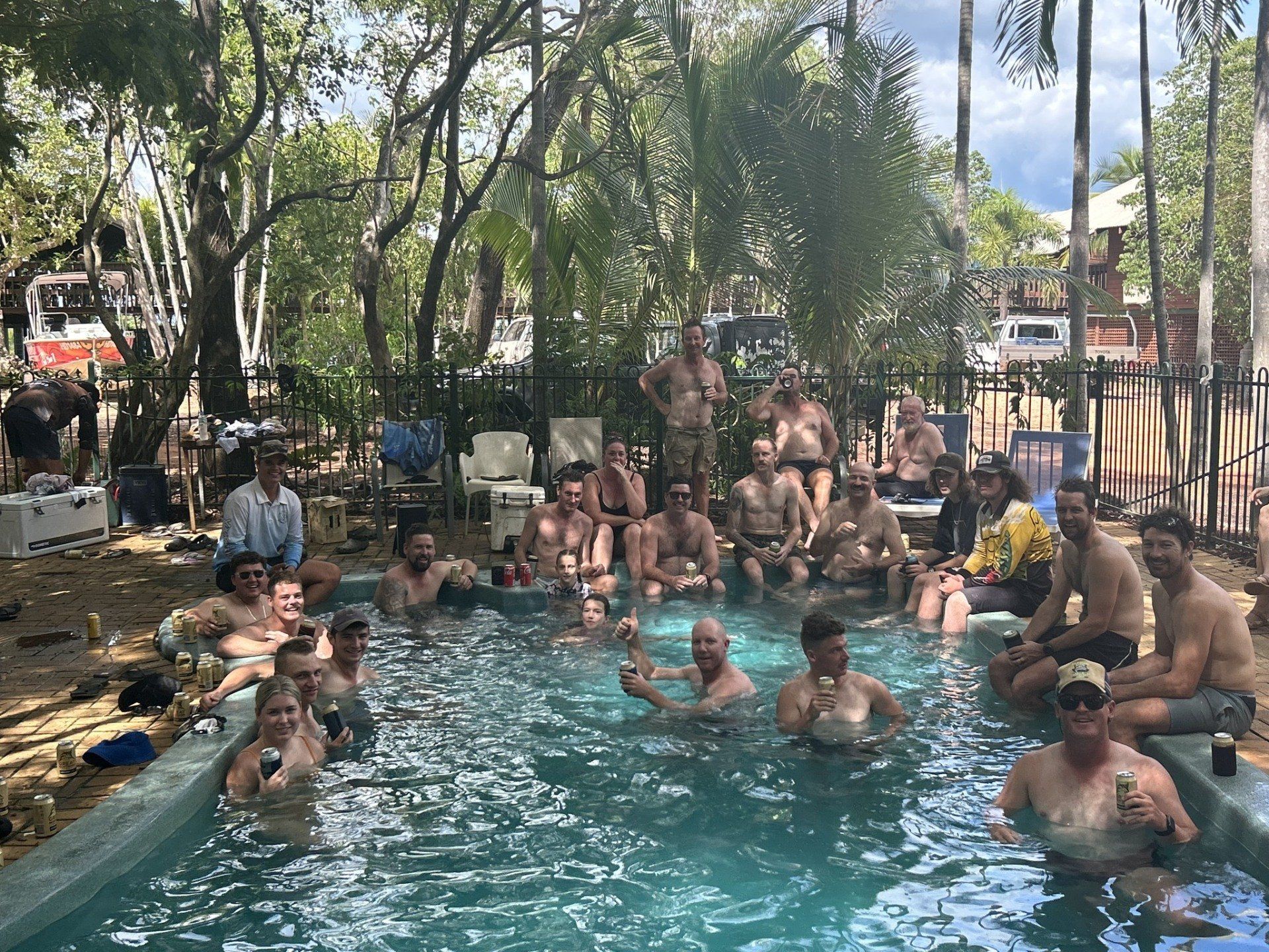 A group of people, some shirtless, relax in a pool outdoors, trees in the background.