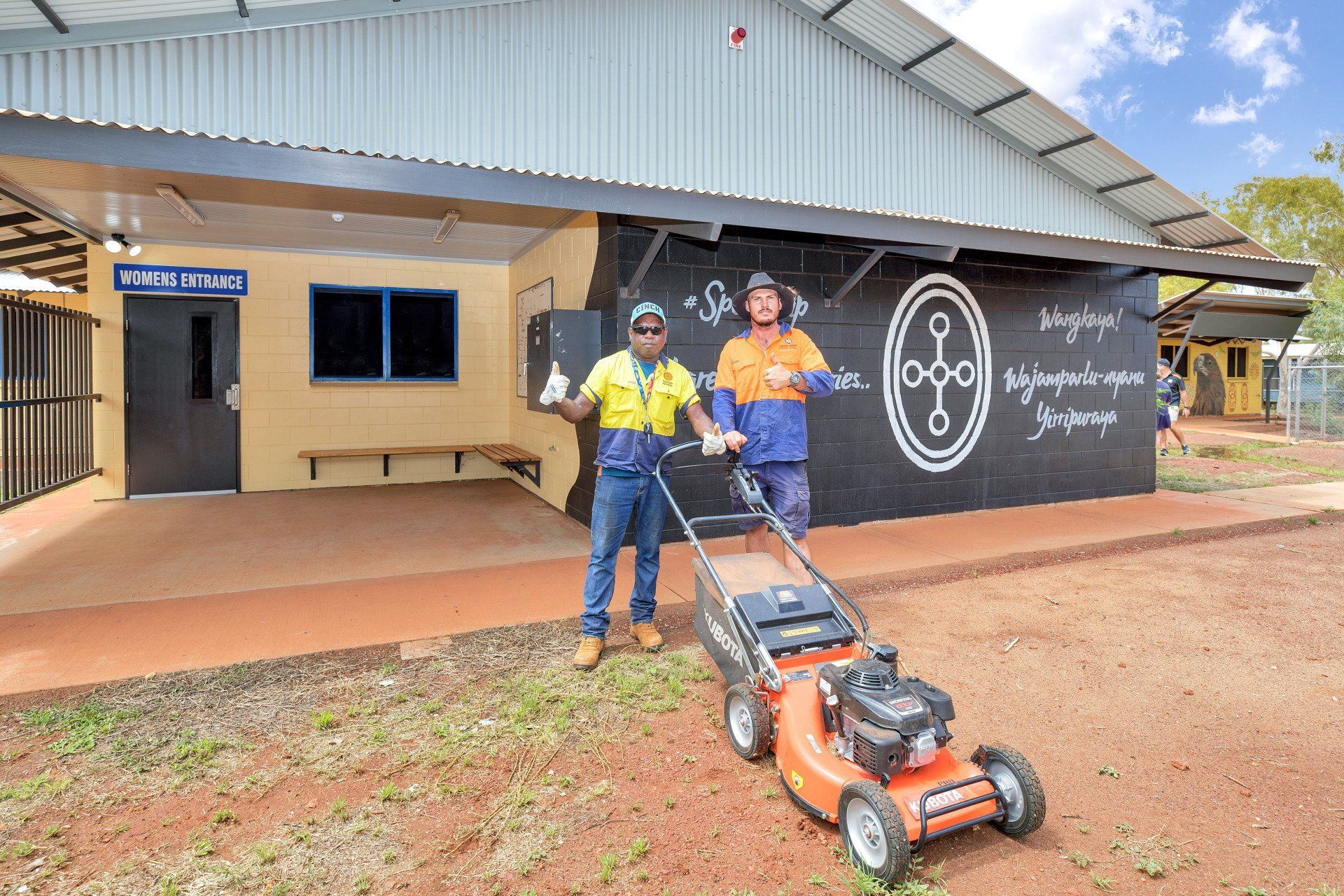 Two men near a building, one mowing the grass. Men wear work clothes. Building has mural and benches.