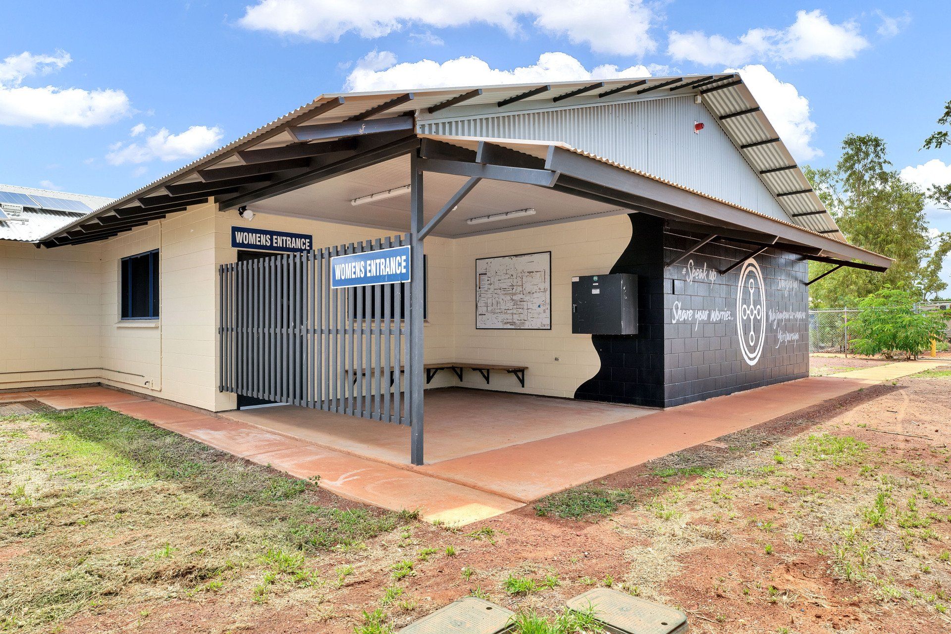 Small beige building with open-air porch, metal roof, and security gate. Blue sky, brown earth.