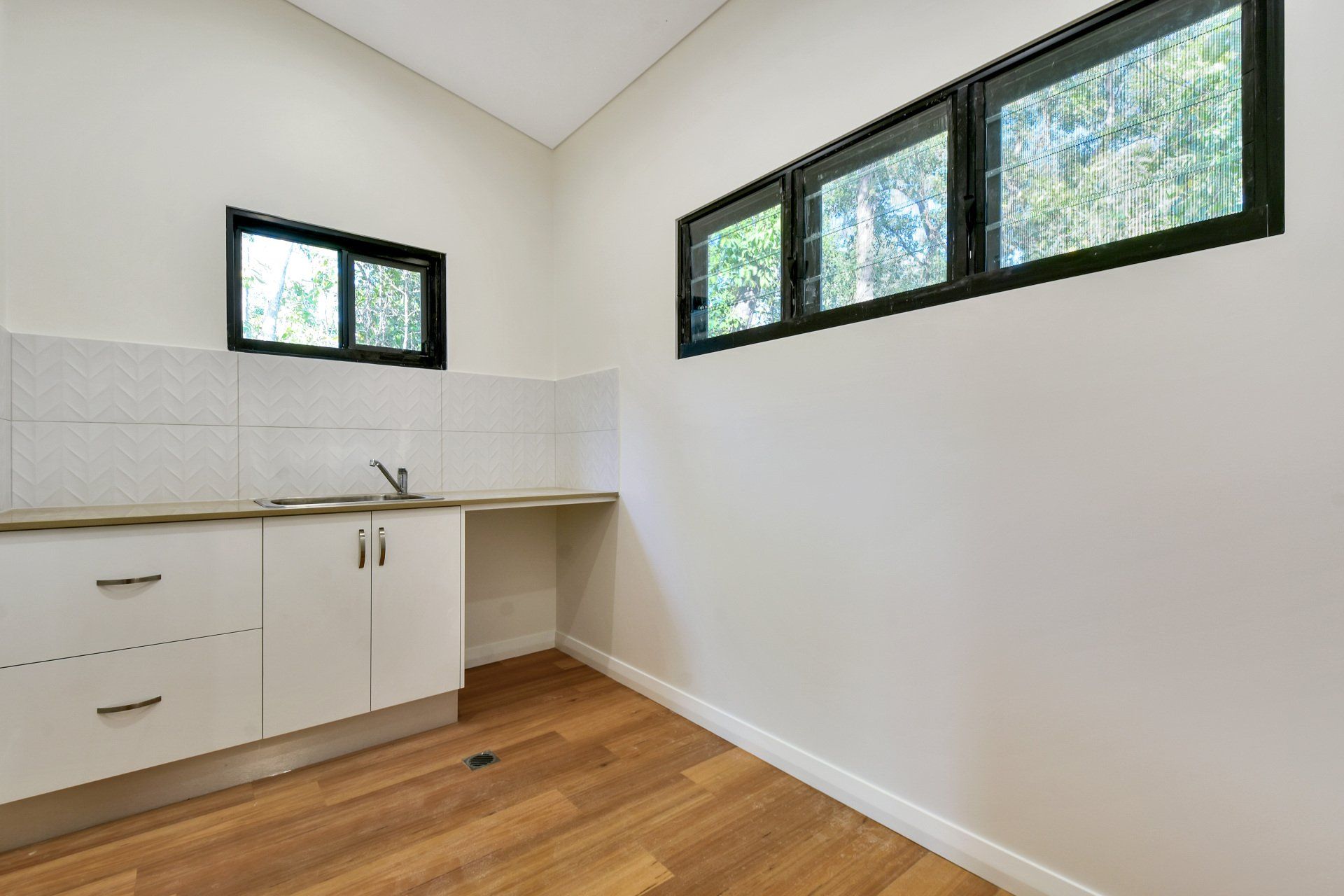 A room with a sink, cabinets, and windows. White walls, wood floor.