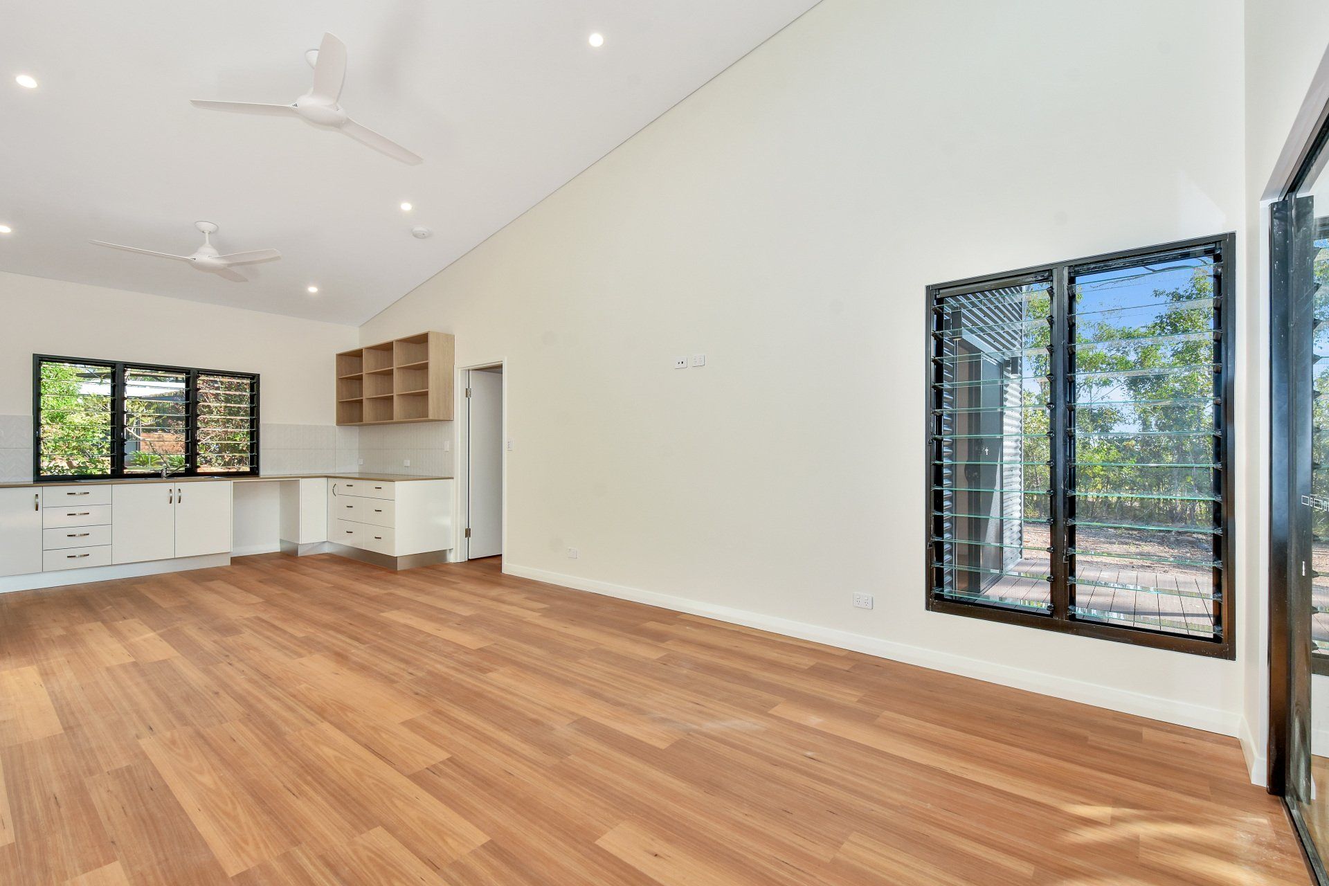 Empty room with wood floor, kitchen, and large windows. Bright, open space.
