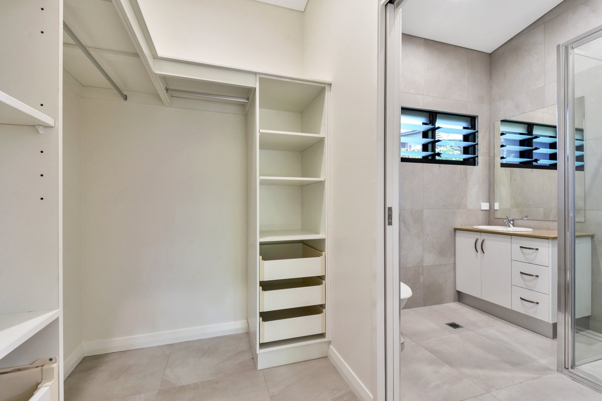 Walk-in closet with shelves, drawers, and hanging rods, leads to a bathroom with a vanity and shower.