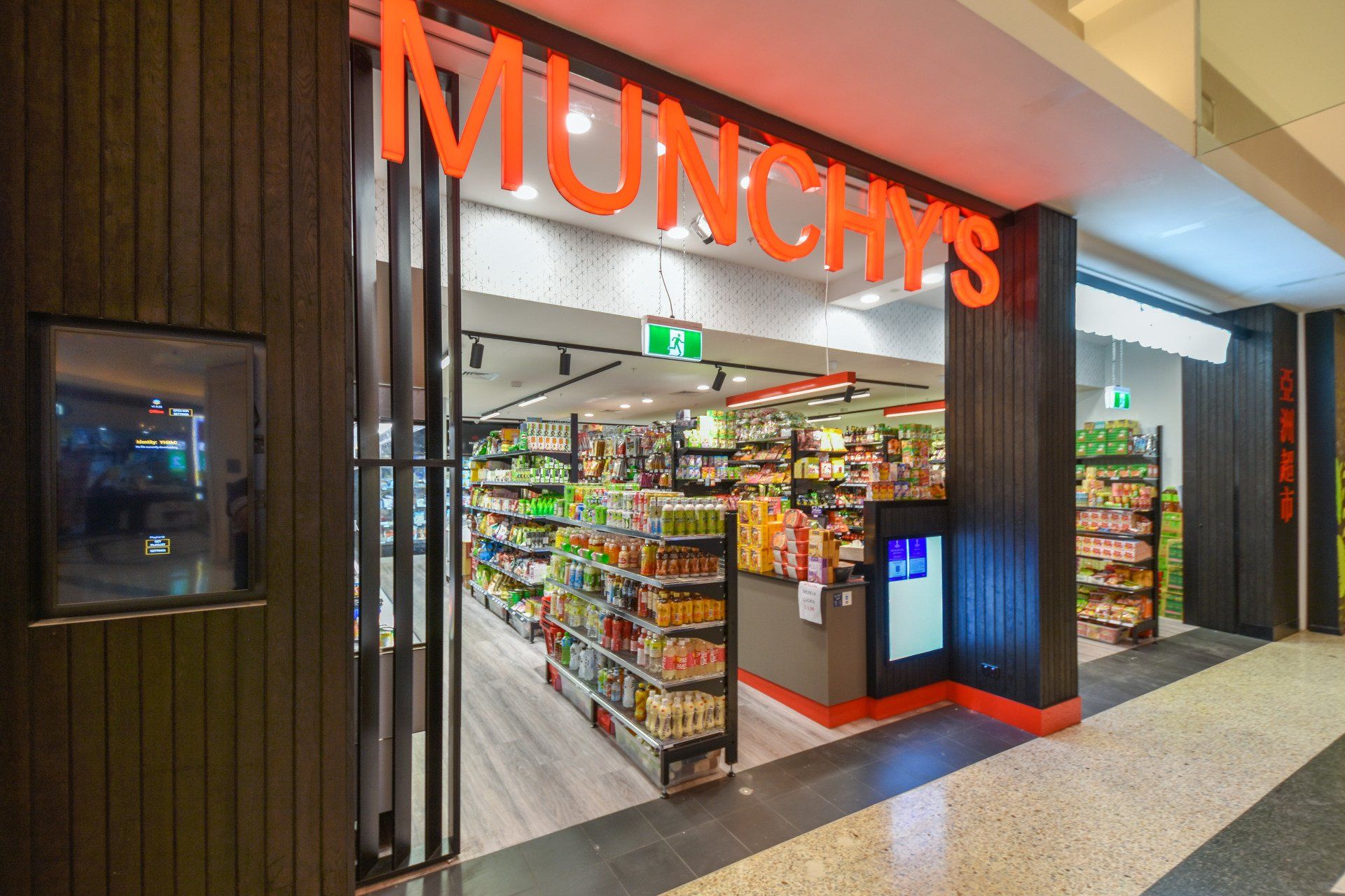 Munchy's store entrance in a mall. Orange sign, wooden facade, bright shelves stocked with food items.