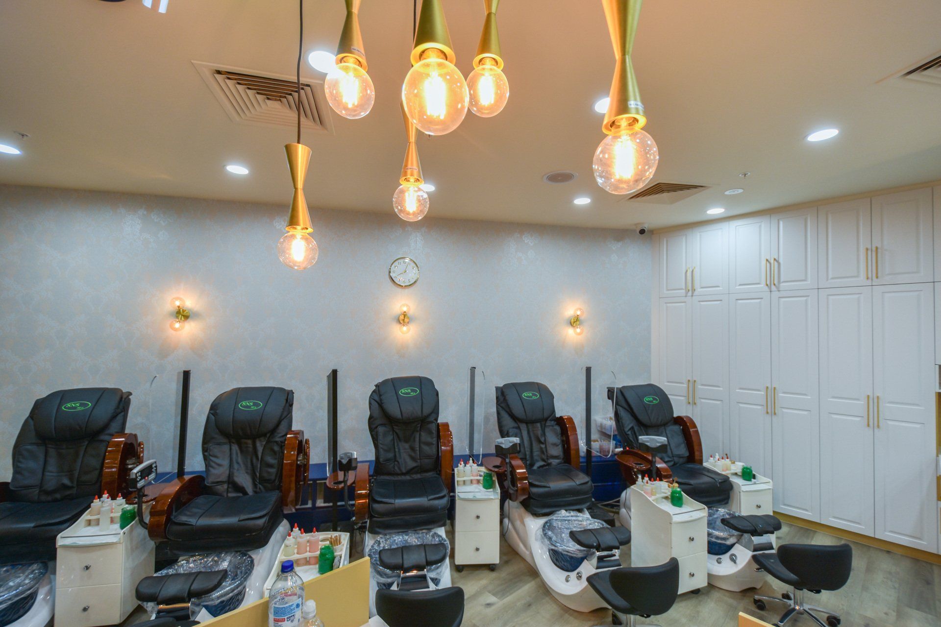 Nail salon with black massage chairs and gold lighting. White storage cabinets on the right.