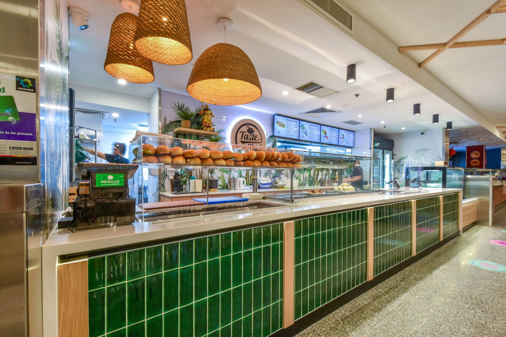 Inside a brightly-lit eatery, a green-tiled counter displays food and menu boards; wicker lamps hang above.