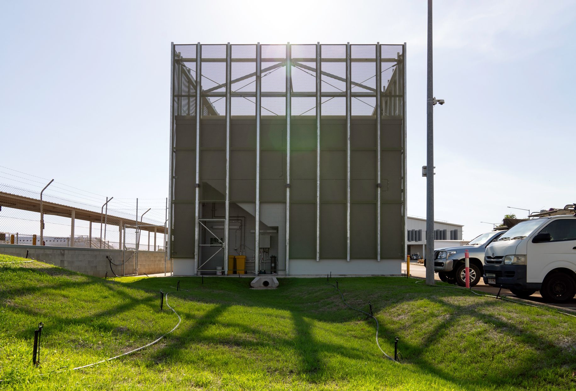 Metal-clad building on grassy hill, with chain-link fence, cars, and a sunny sky.