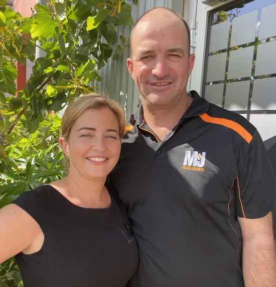 Woman and man smiling, standing close outdoors in front of a building and green leaves. Man wearing a black shirt.