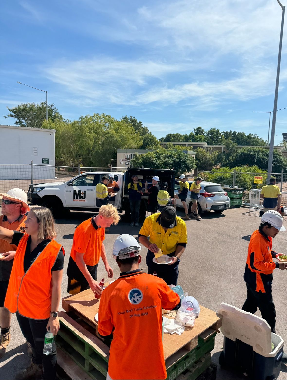 Construction workers in orange vests and helmets at a lunch break outdoors, sunny sky.