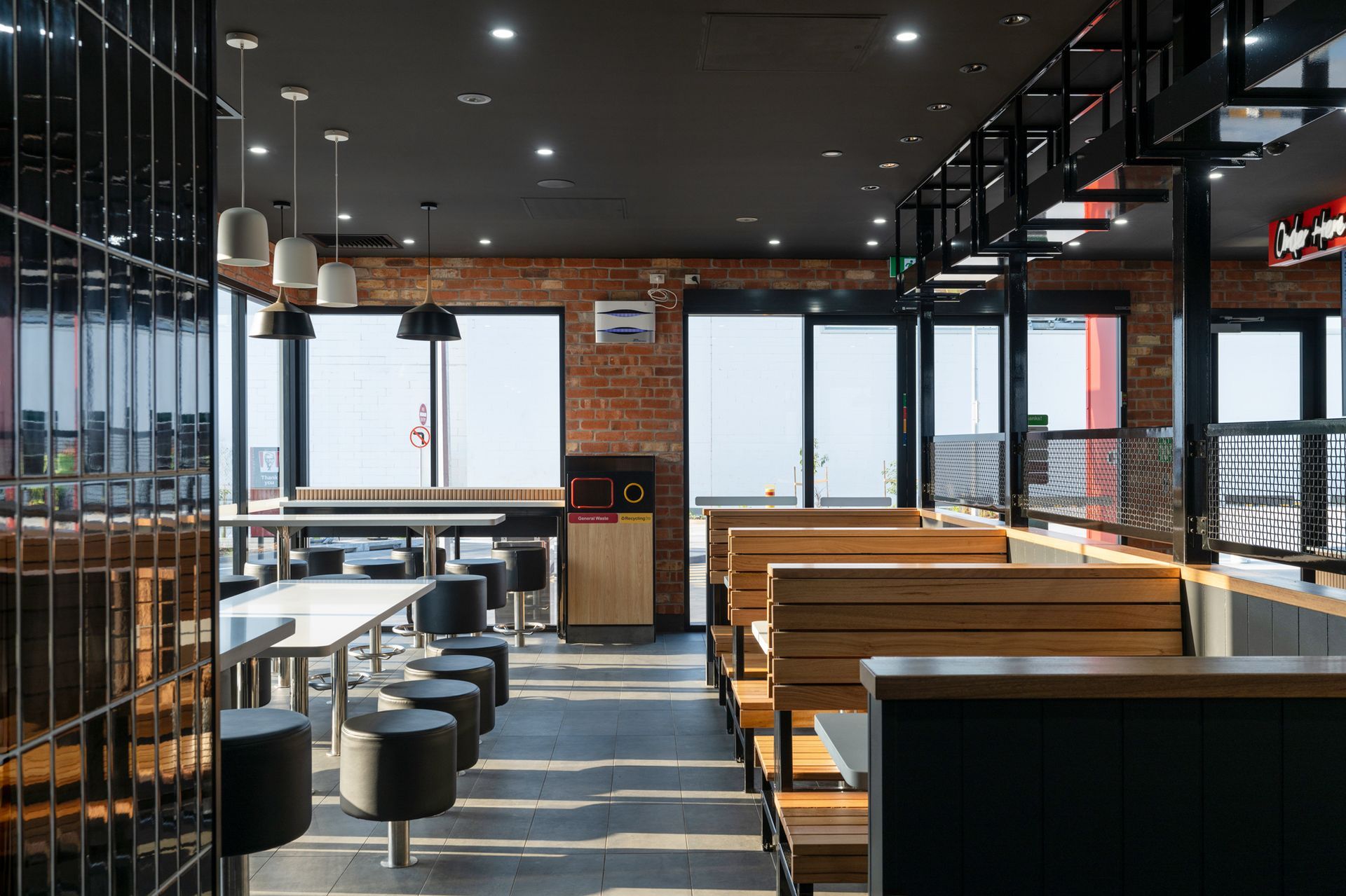 Interior of a modern restaurant with brick walls, tables, stools, and booths, with large windows offering a view.