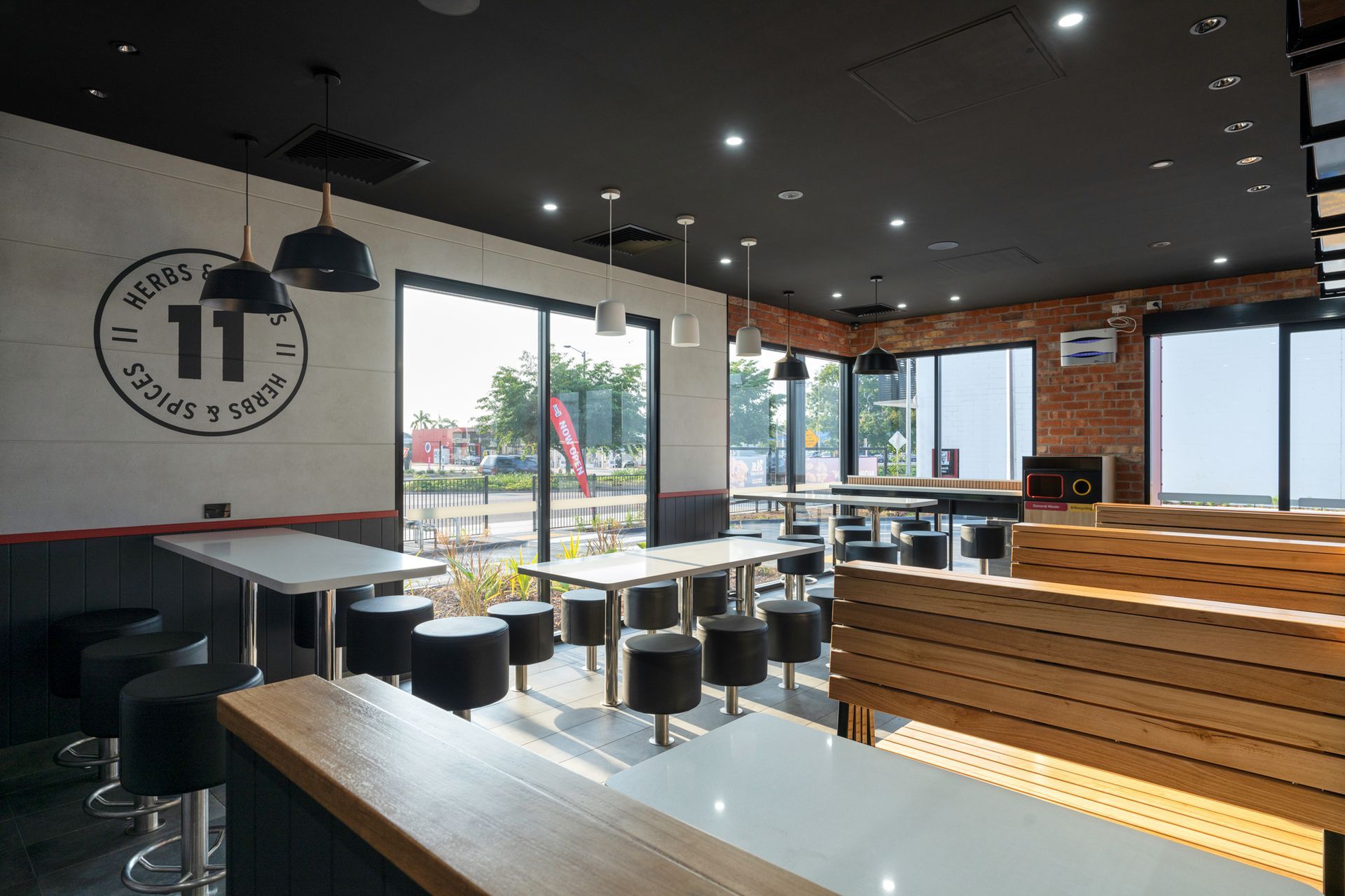 Interior of a restaurant with tables, stools, and large windows; black ceiling, brick wall, and logo on the wall.