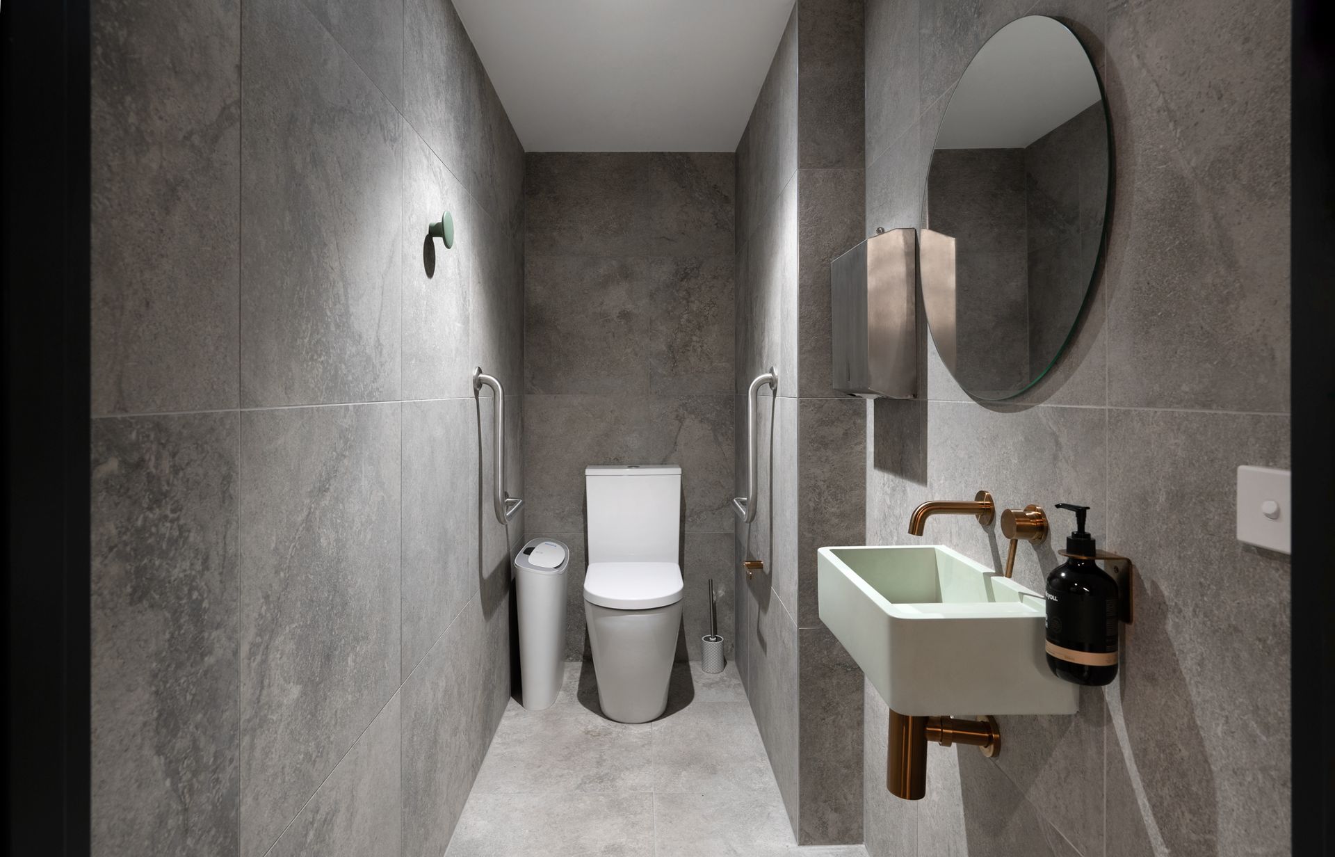 Modern gray tiled bathroom with toilet, sink, and round mirror.