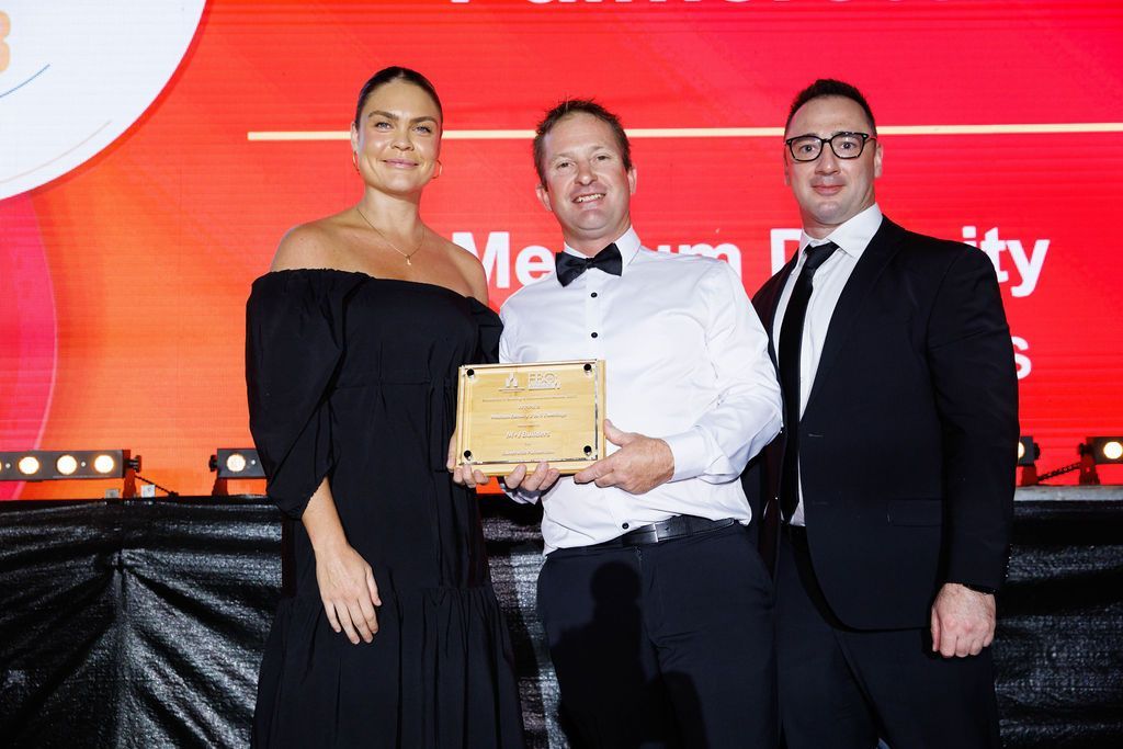 Three people on stage, one holding a plaque. Woman in black dress, two men in formal wear. Red background.