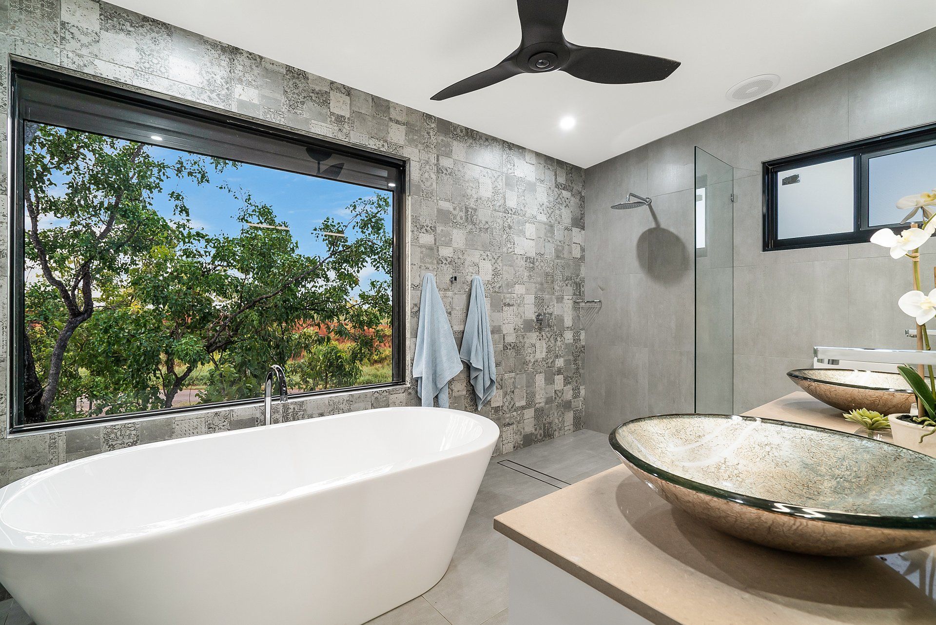 Residental bathroom with his and her vanities, waterfall showerhead bath below a large window looking out to a tree.