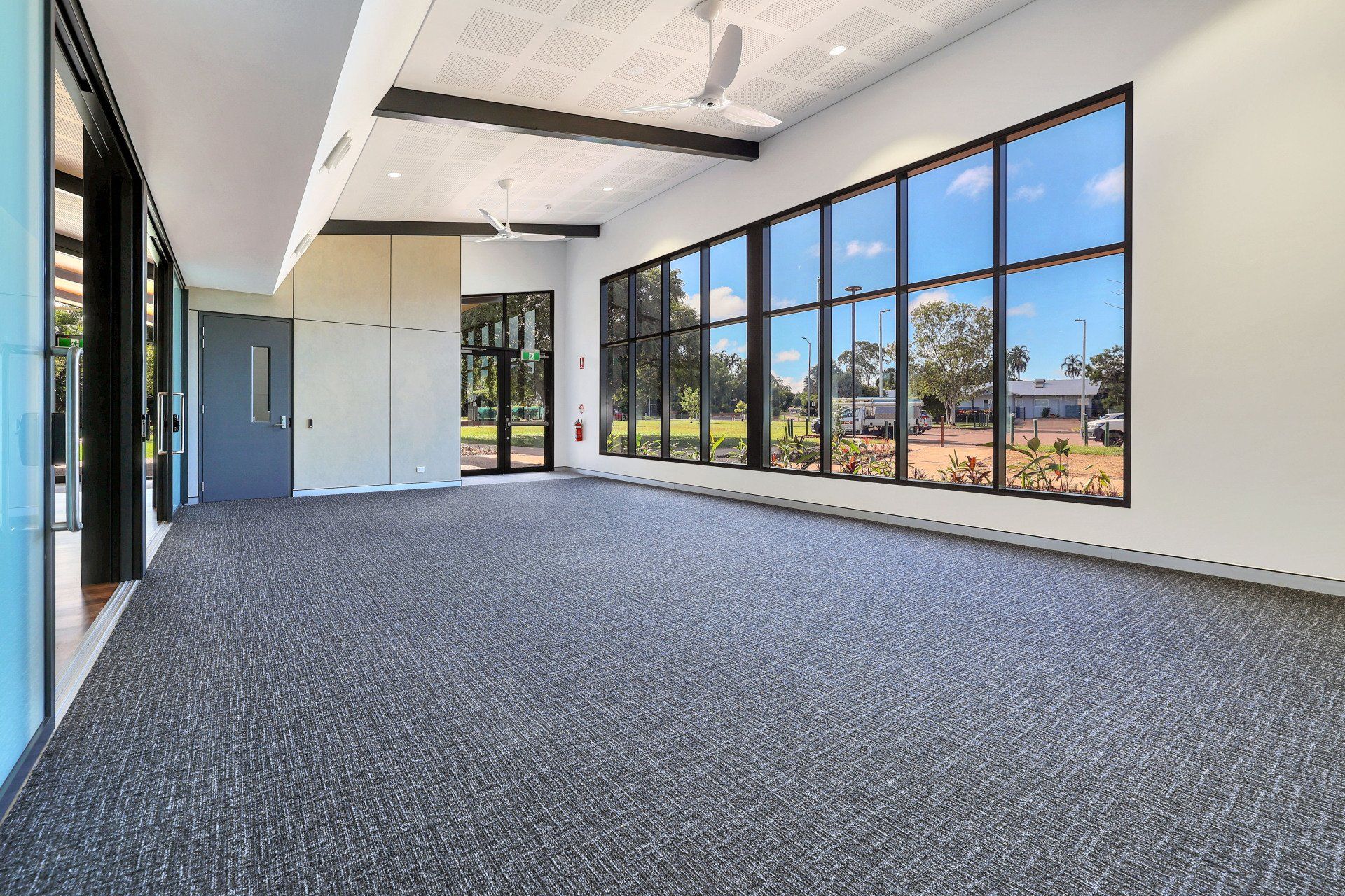 Empty room with large windows overlooking a green space; gray carpet, white walls, and ceiling.