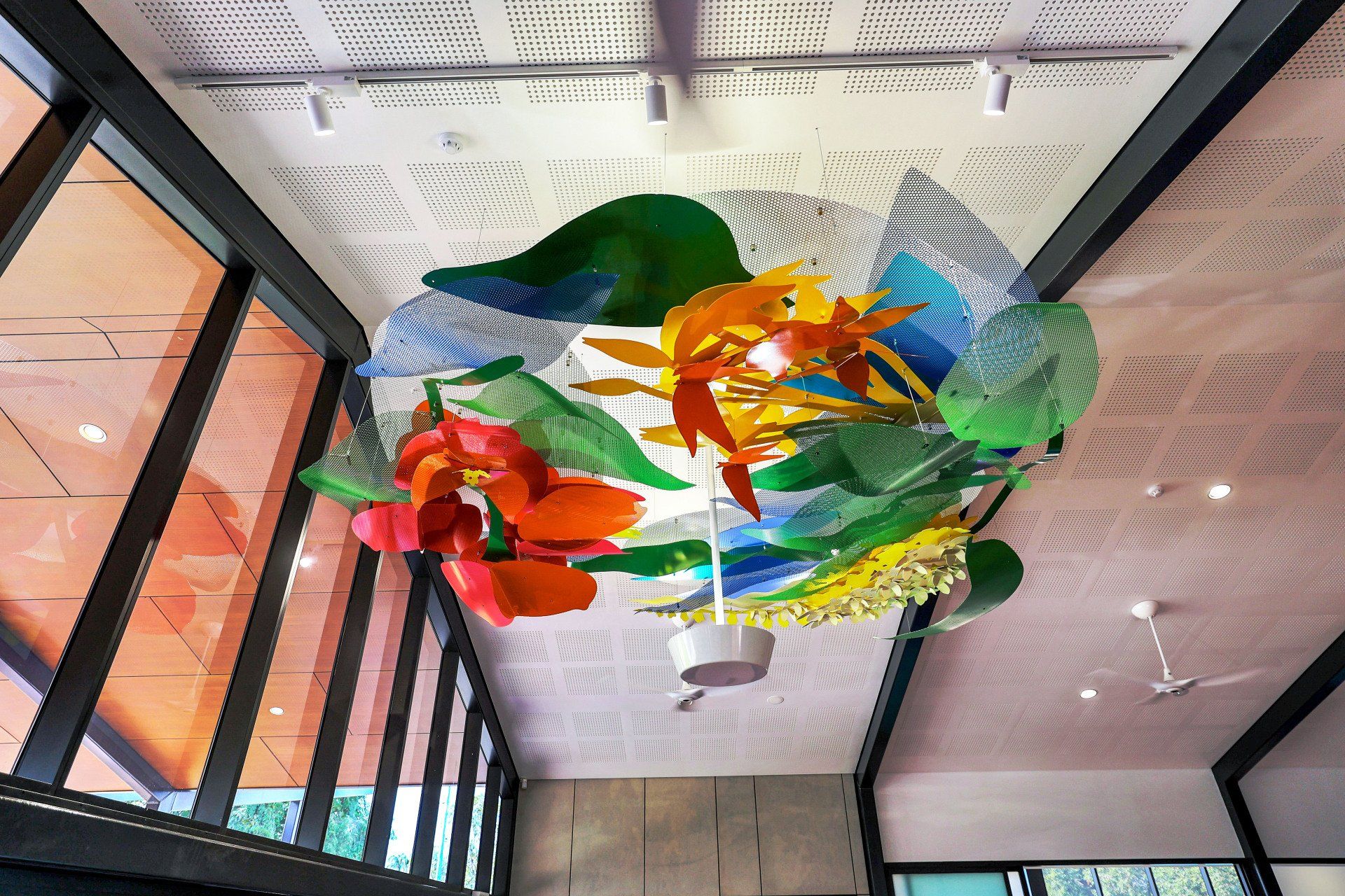 Colorful glass art installation suspended from the ceiling. Features leaves and flowers in a room with windows.