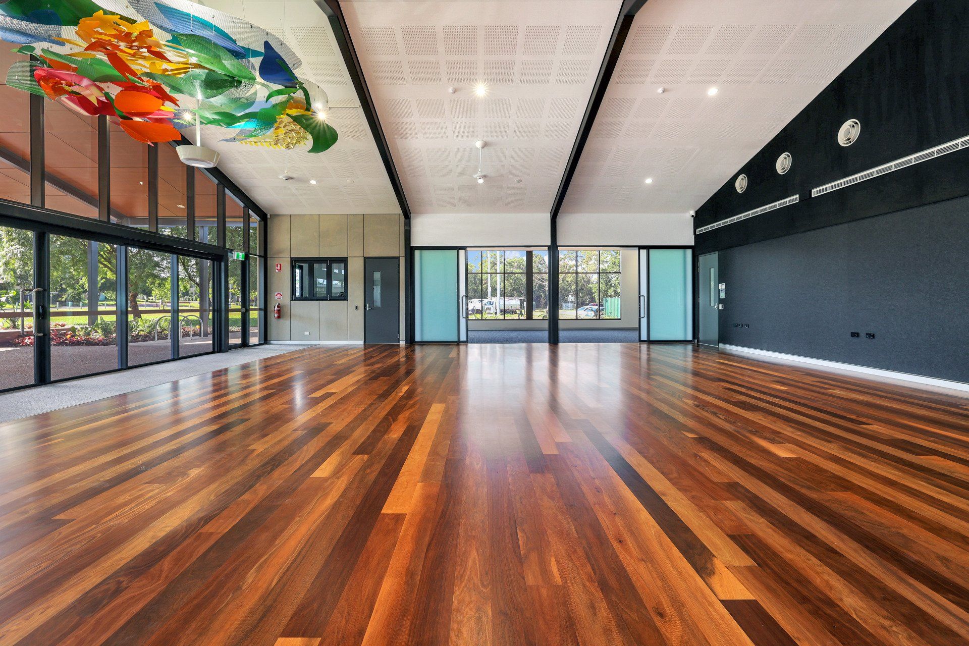 Large, empty hall with wooden floor, glass walls, and colorful ceiling art.