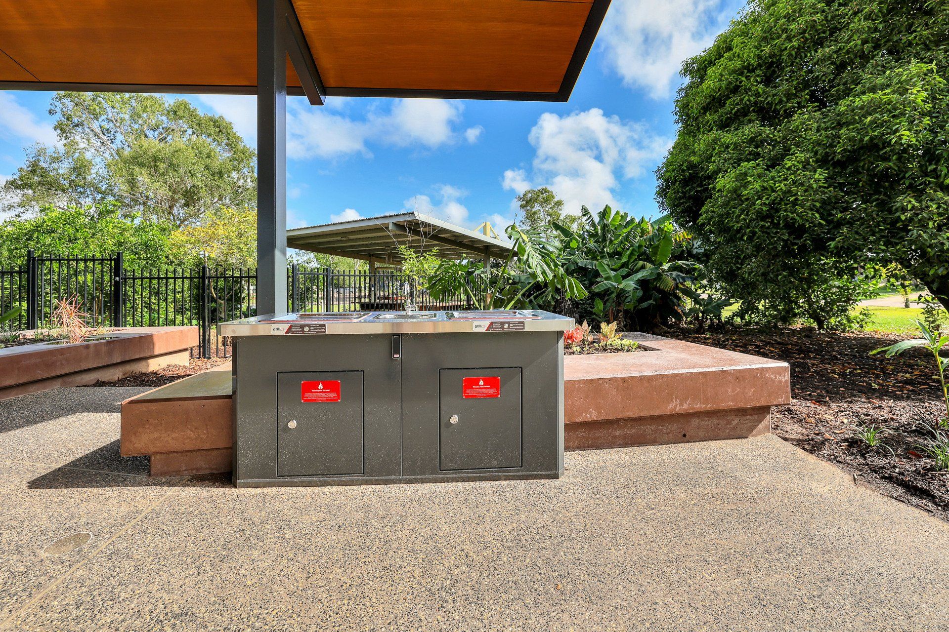Barbecue grill under a wooden roof in a park setting, surrounded by greenery, blue sky, and concrete.