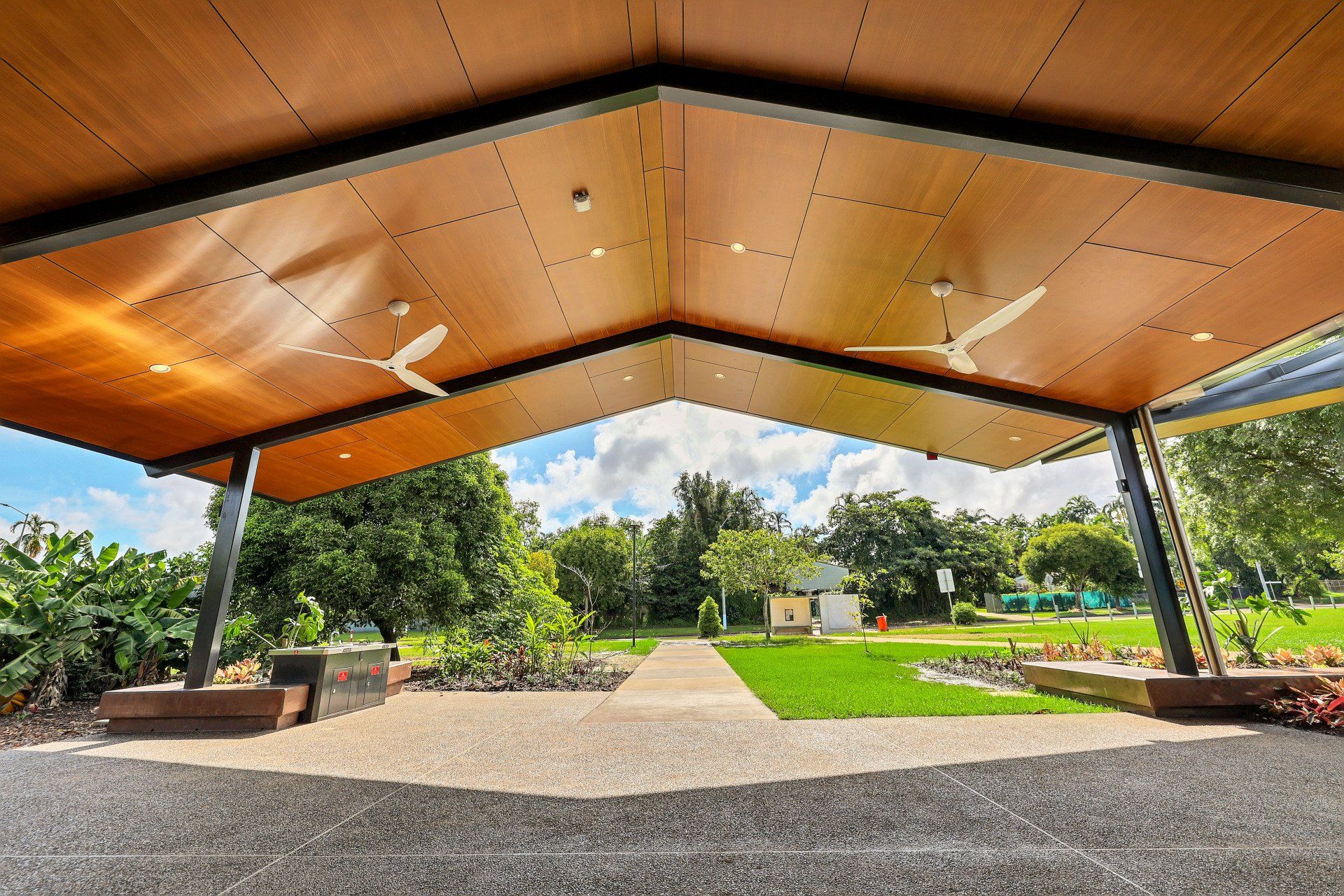 Patio with a wood ceiling, overlooking a green lawn with trees and a small building under a blue sky.