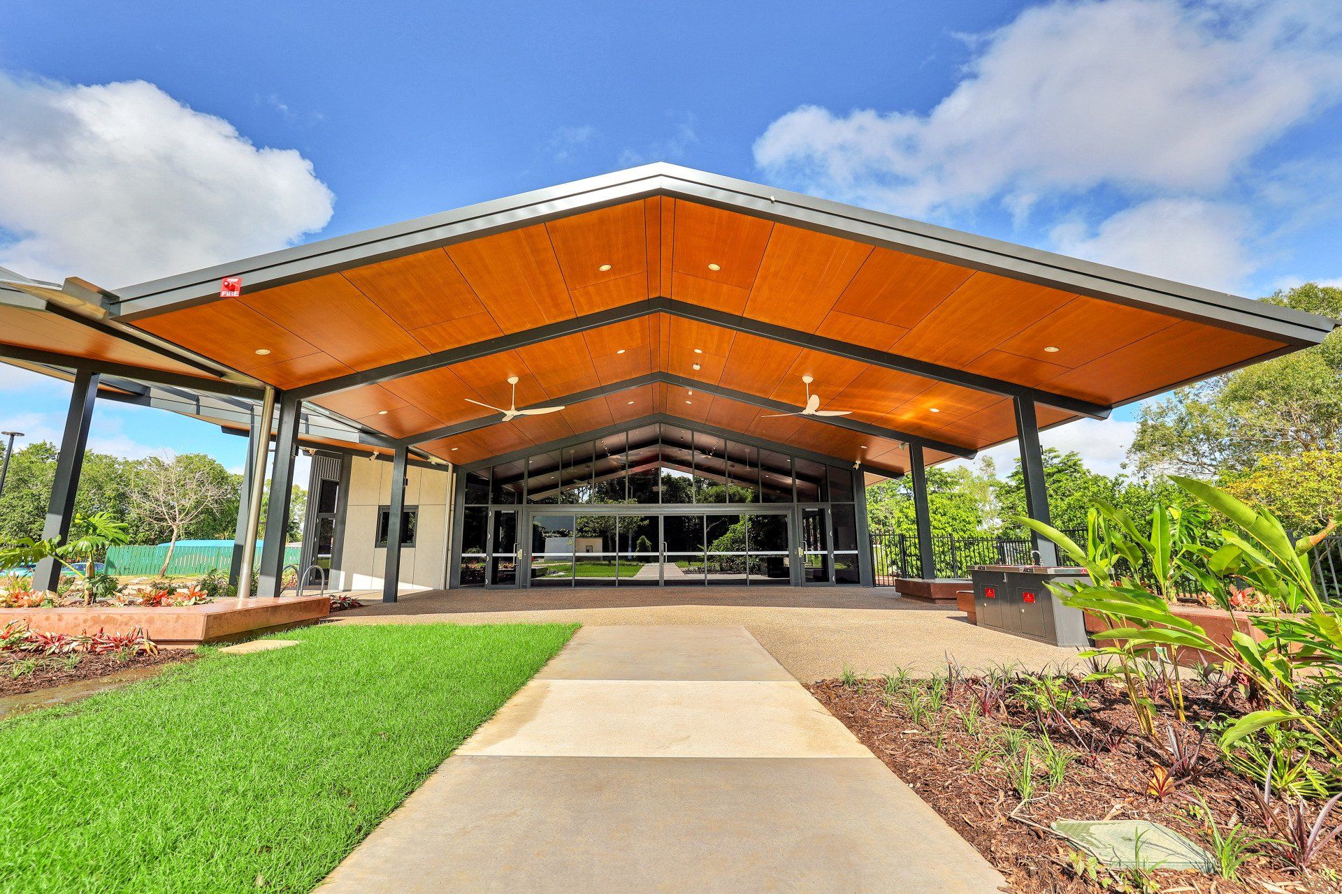 Entrance to a modern building with a sloped wooden ceiling, glass doors, and a paved walkway on a sunny day.