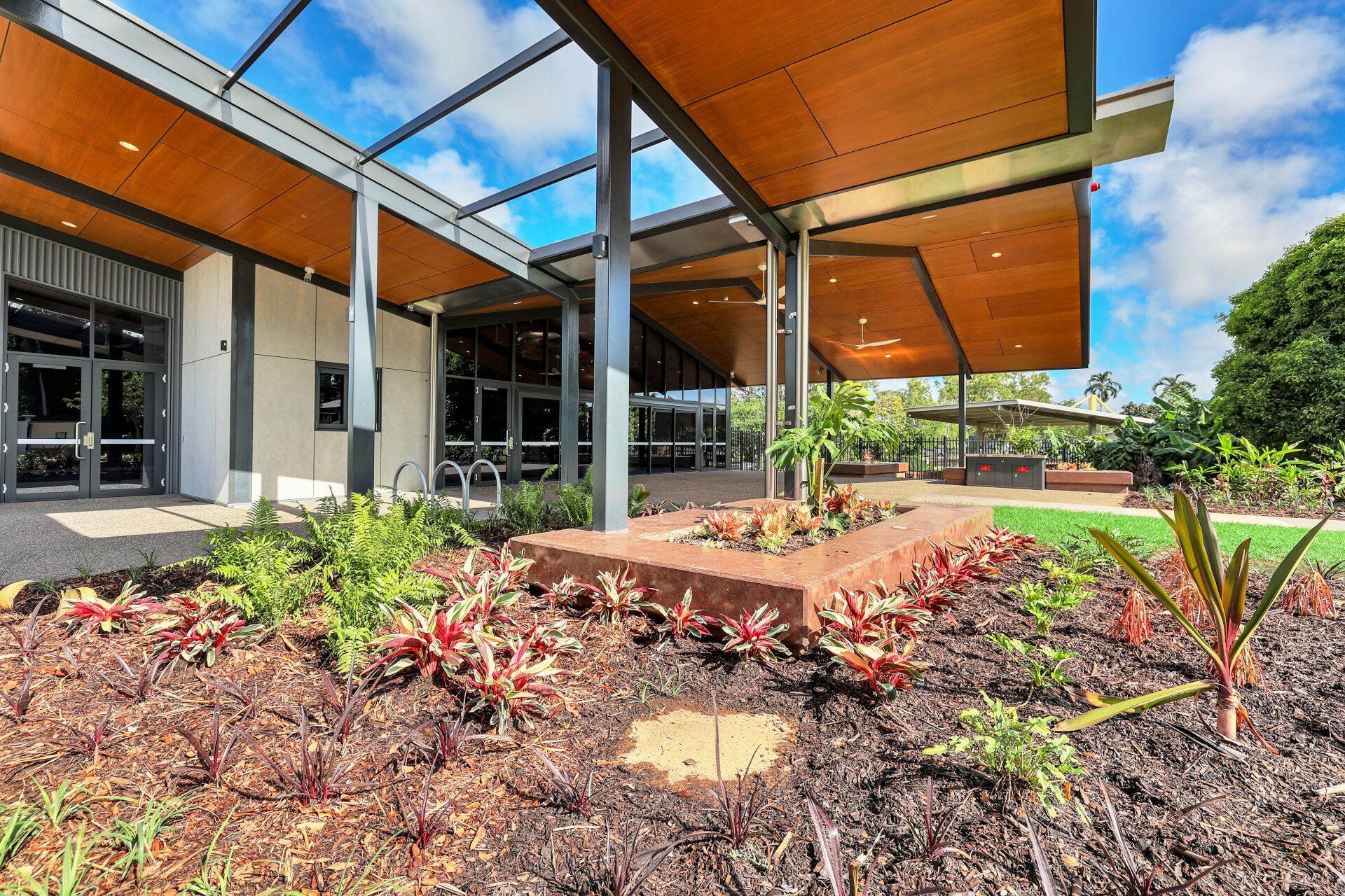 Building entrance with brown wooden ceiling, glass doors, landscaped garden with colorful plants.