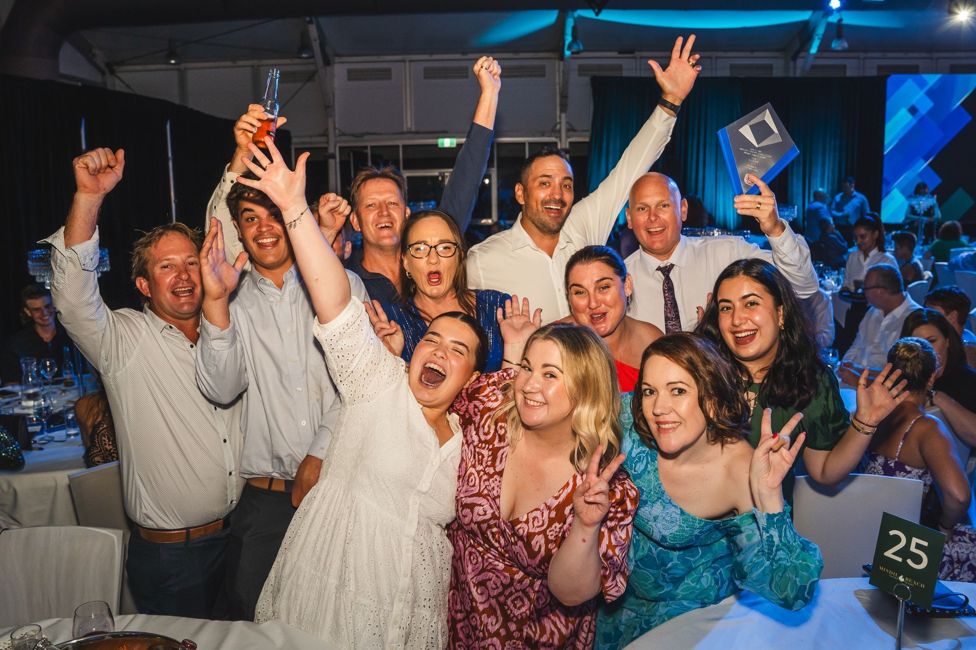 Group of people celebrating at an event, arms raised, excited expressions, with a trophy and champagne.