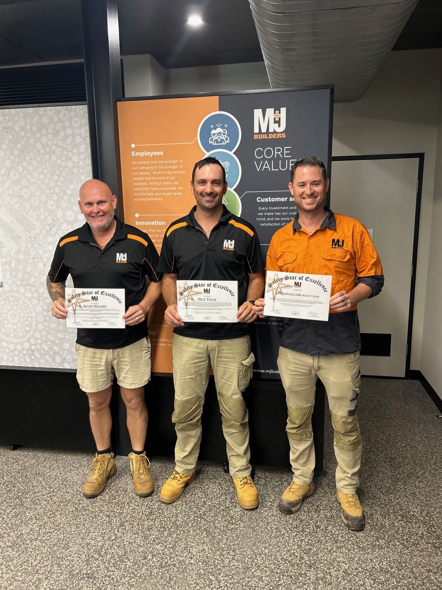 Three men in work clothes holding certificates, standing in front of a sign; indoor setting.