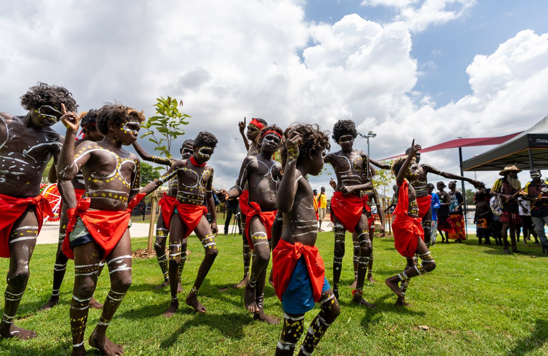 Indigenous Australians in red skirts and body paint dancing on a grassy field under a cloudy sky.