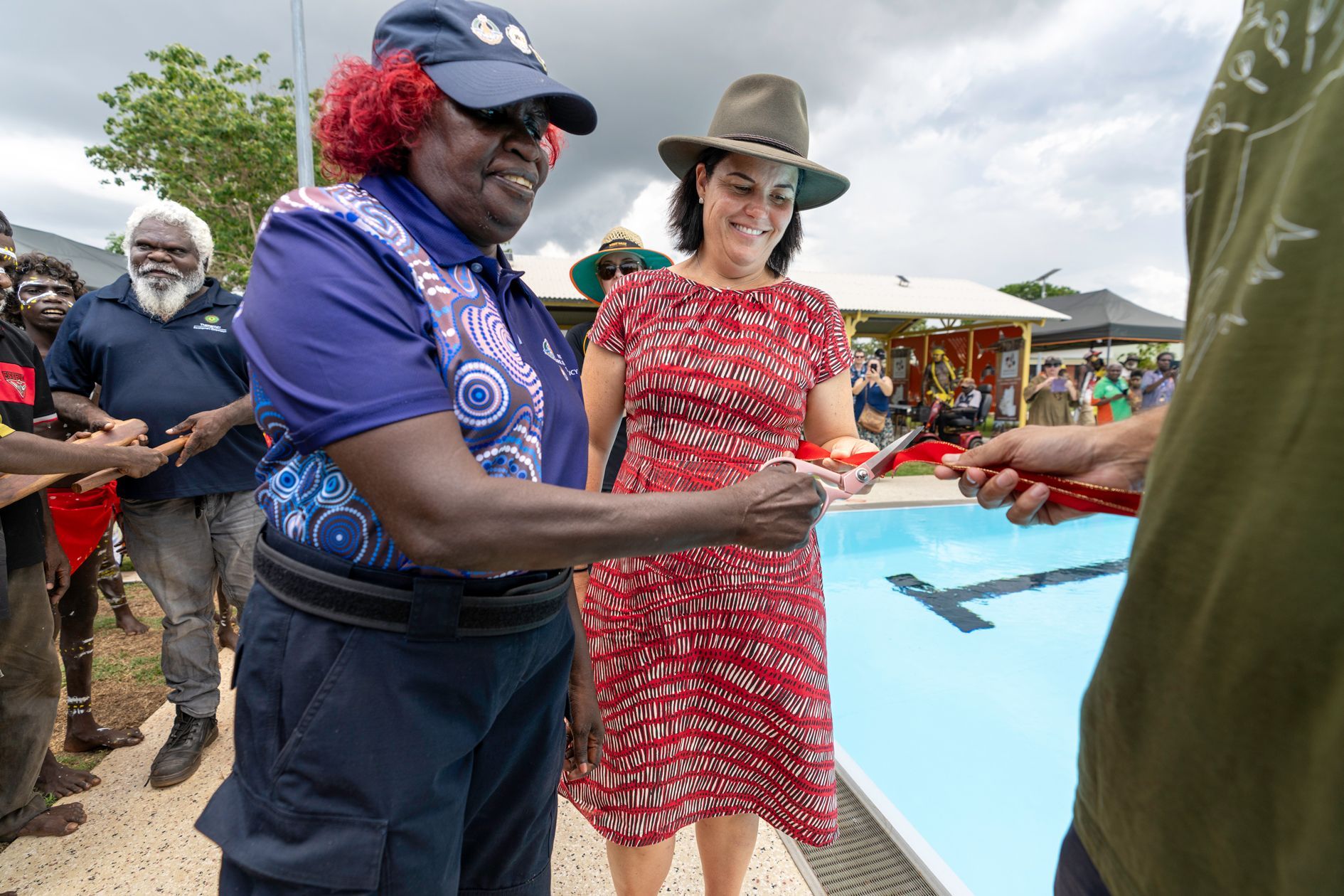 Two women cut a ribbon at a pool opening. Indigenous woman in blue, another in red dress. Crowd watches.