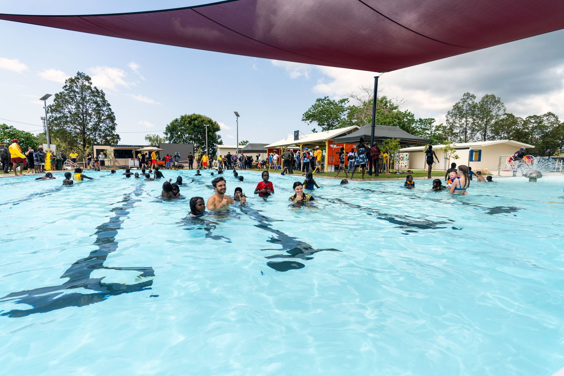 People swimming in a blue outdoor pool under a shade canopy on a sunny day.
