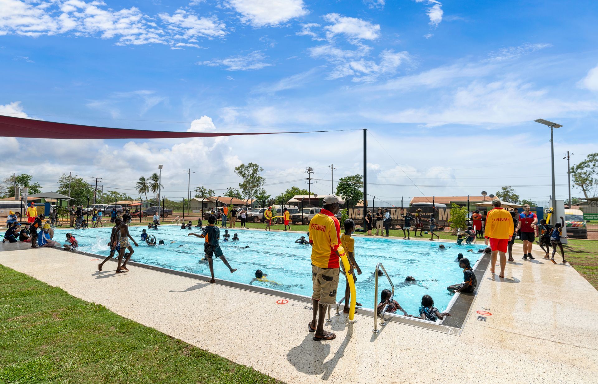 People swimming in a public pool on a sunny day, with lifeguards on duty and shade.