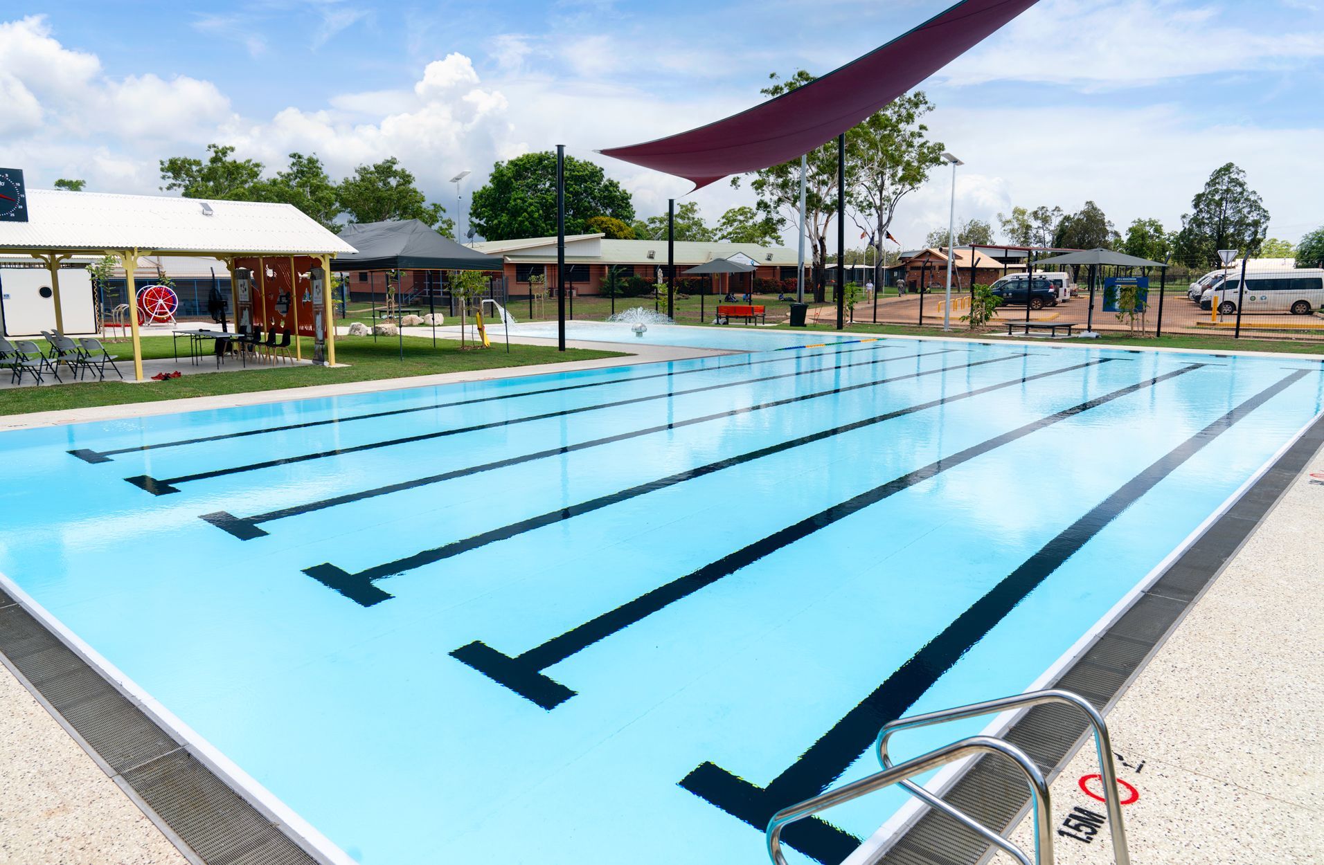 A rectangular swimming pool with lane markers, under a blue sky. Red shade sail in the background.