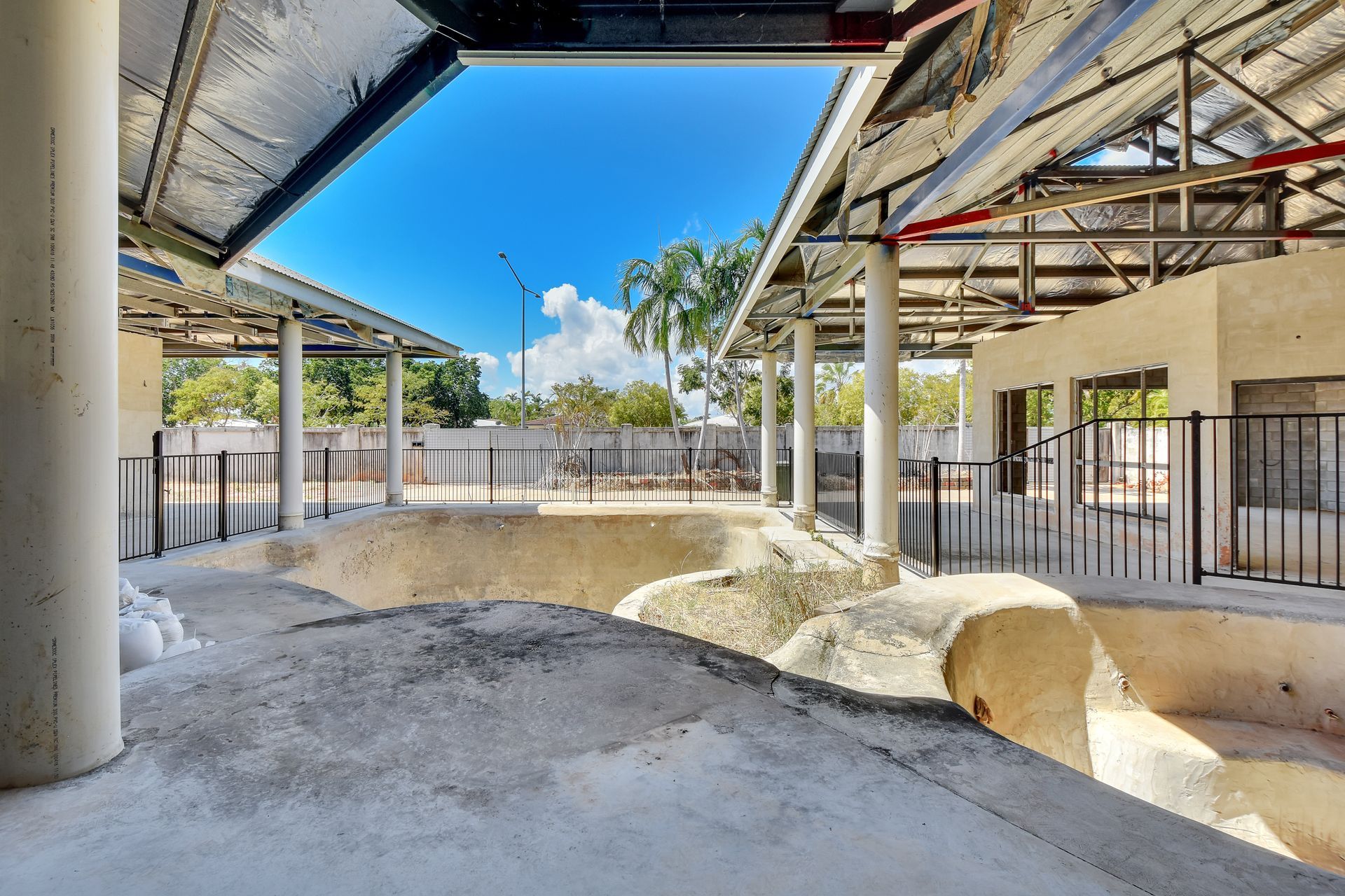 Construction site, open-air structure under a bright blue sky. Concrete floors, pillars, and metal framing are visible.