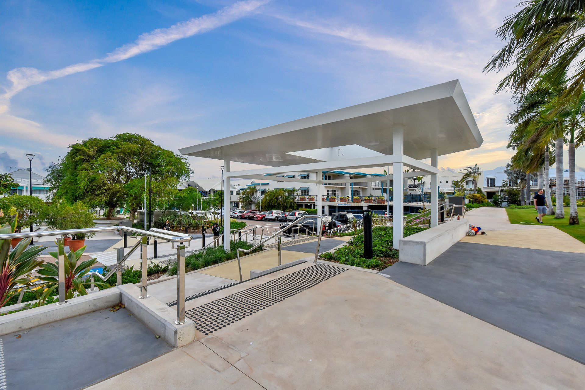 Park with white shelter and stairs, pathway, trees, and waterfront buildings under a blue sky.
