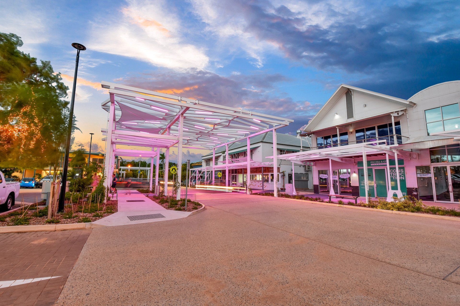 Entrance to a modern building with a white pergola illuminated by pink light, under a colorful sunset sky.