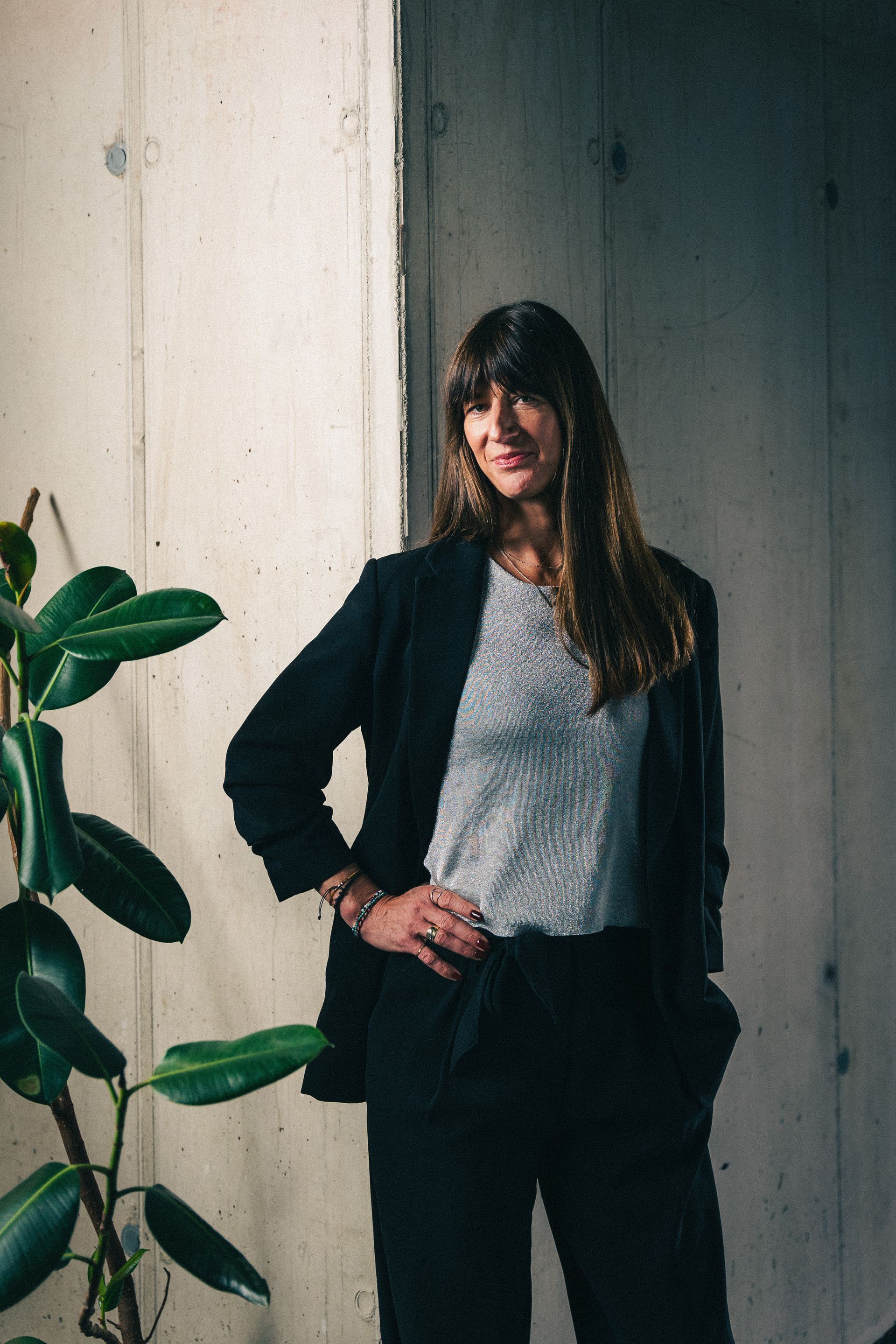 Person standing indoors beside a plant, wearing a black blazer and gray top against a light wall.